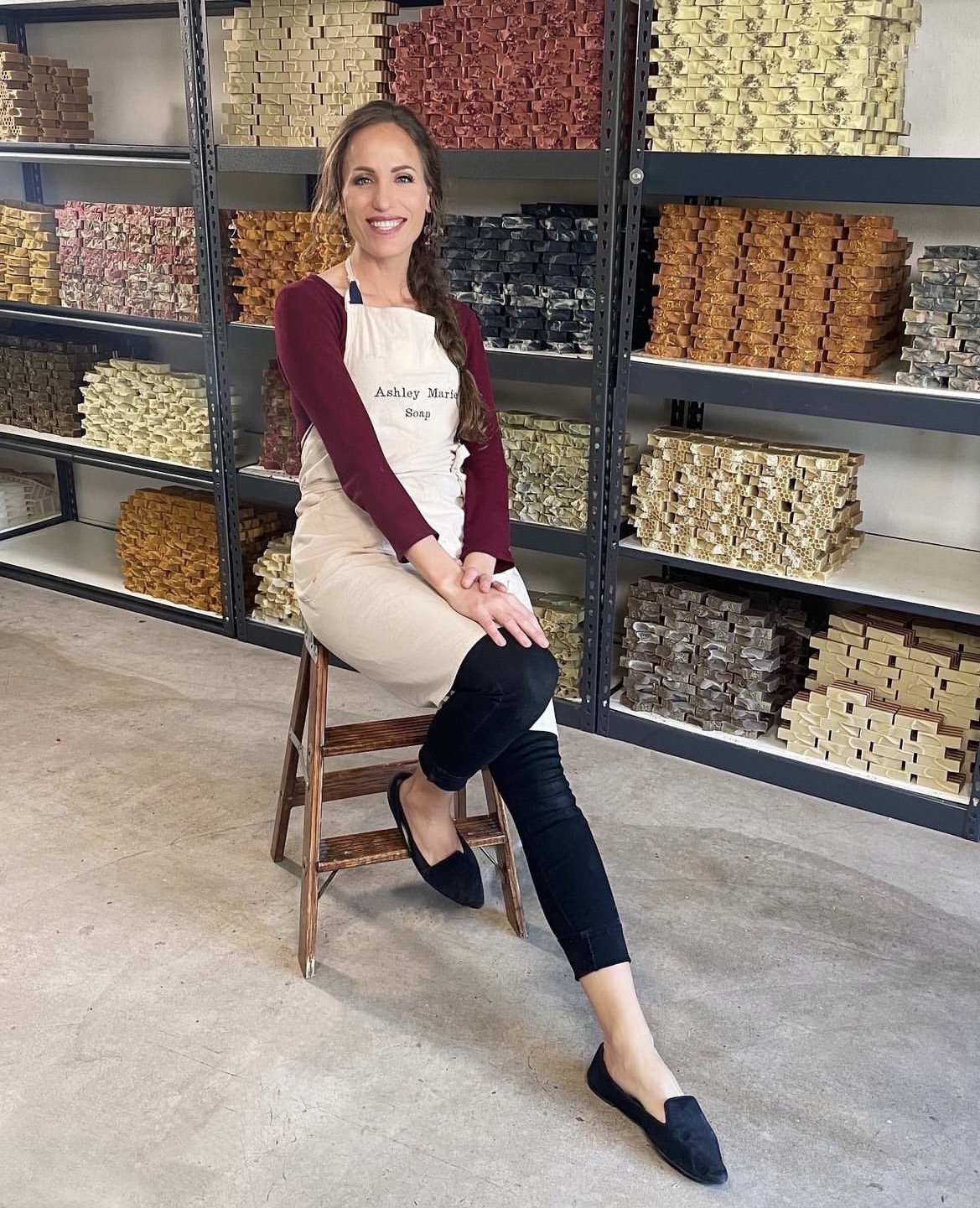 Woman sitting on a stool in front of shelves filled with colorful boxes, smiling, wearing a beige apron and dark pants.