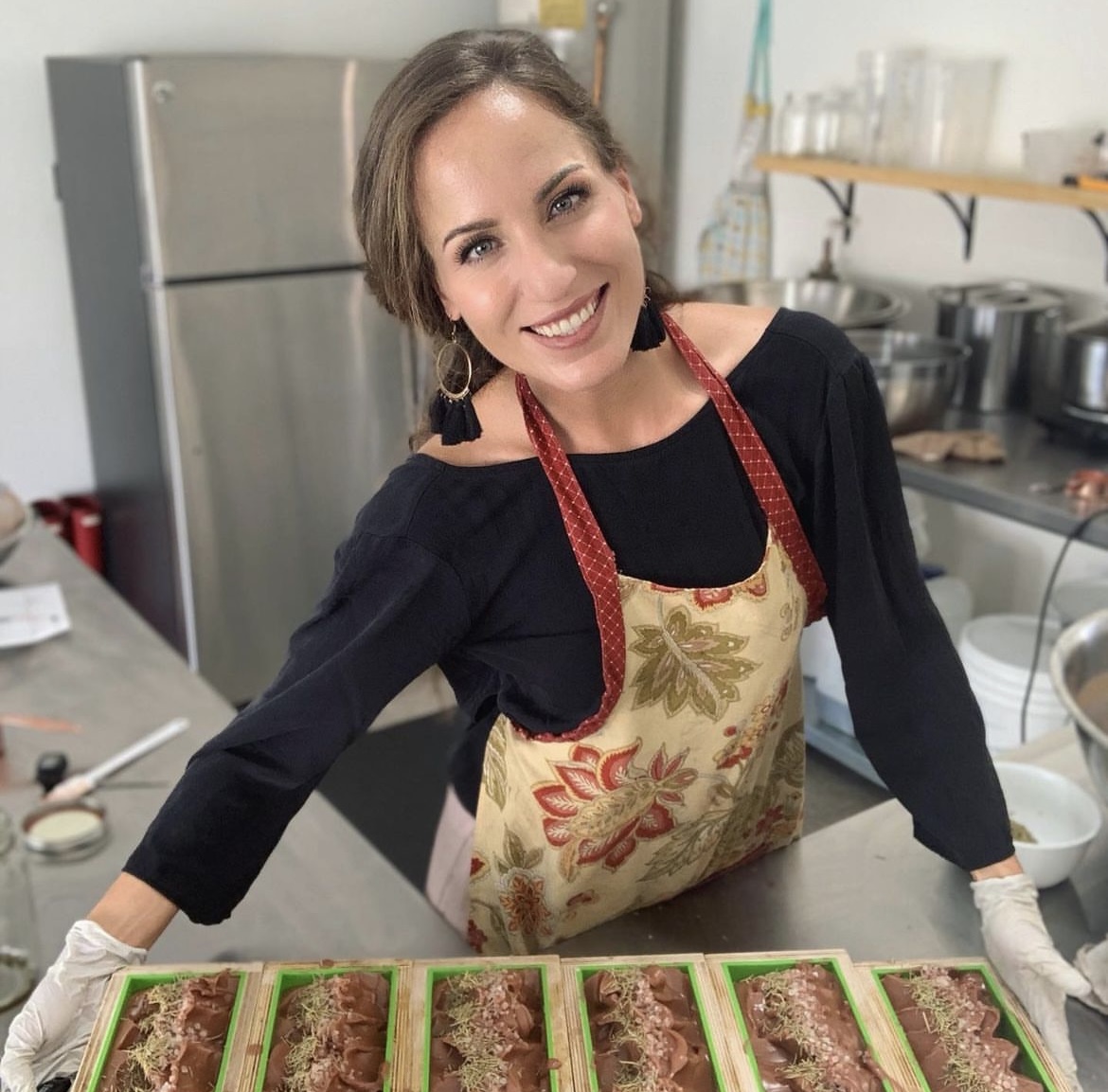Woman smiling in a kitchen, wearing an apron, standing behind a tray of food, with kitchen appliances in the background.