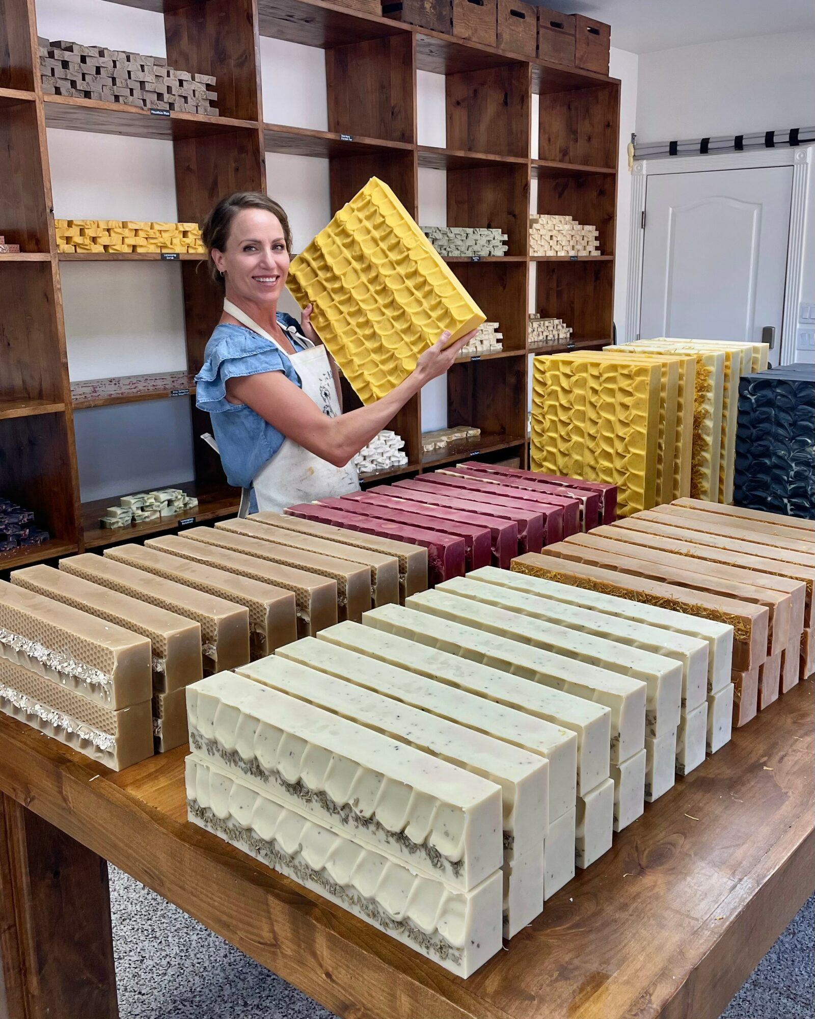 Woman holding a yellow soap mold in a room with shelves and soap bars on a table.