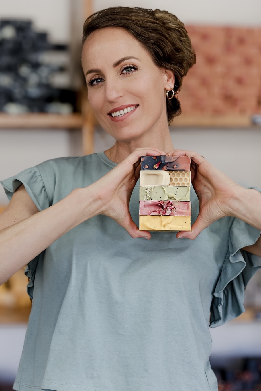 Woman smiling holding a stack of colorful gift boxes with a heart shape, in a room with shelves in background.