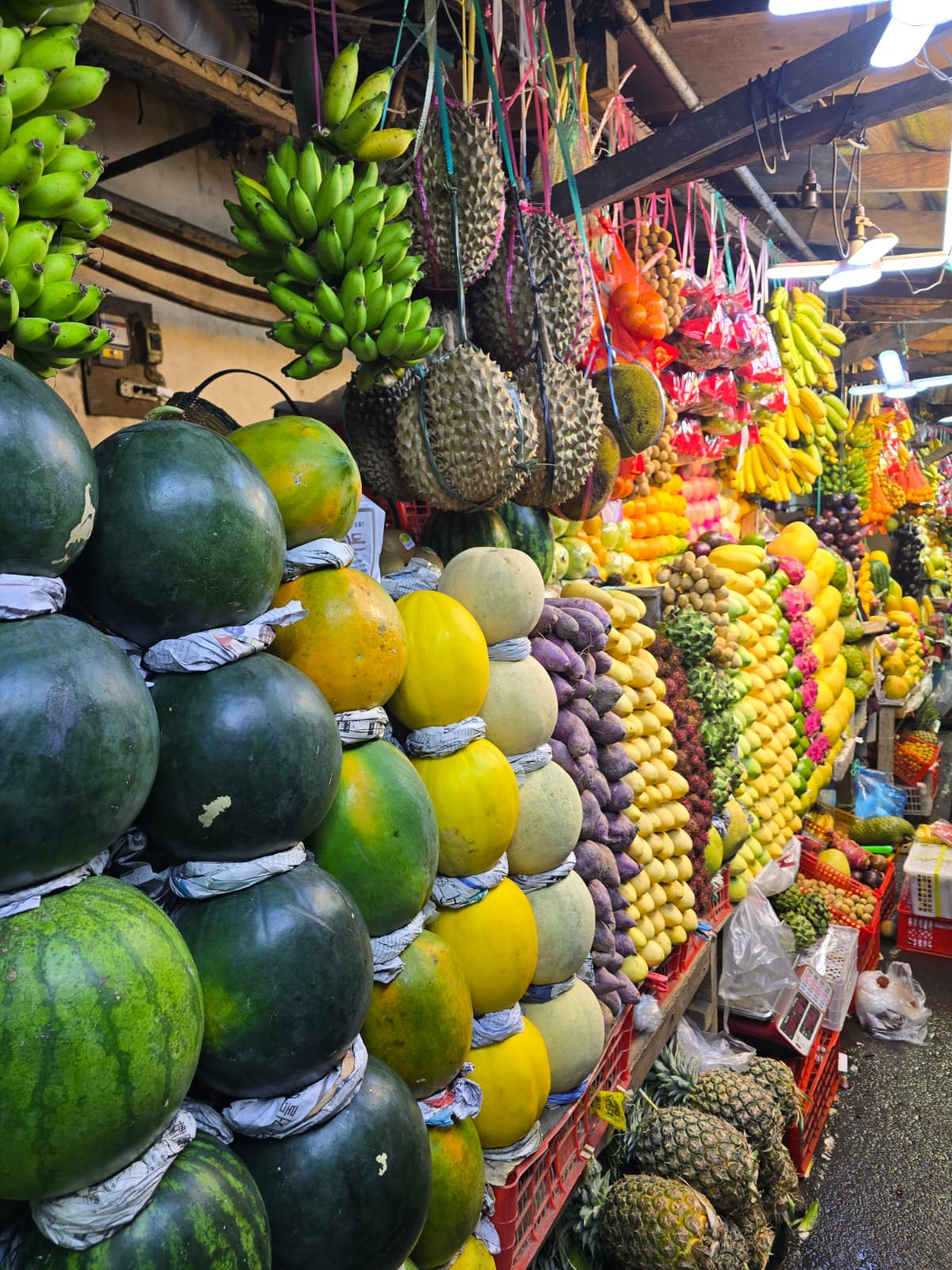 Display of various fruits including watermelons, bananas, lemons, and pineapples at a market stall.