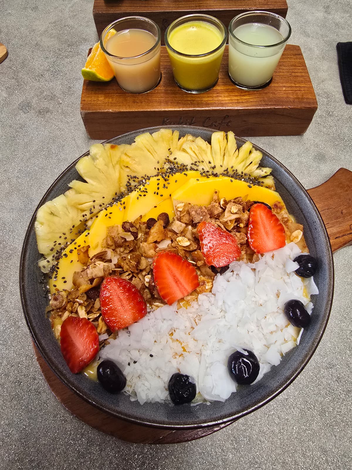 Bowl of rice with strawberries, pineapple, and black olives, alongside a wooden tray with three small drinks in glasses.