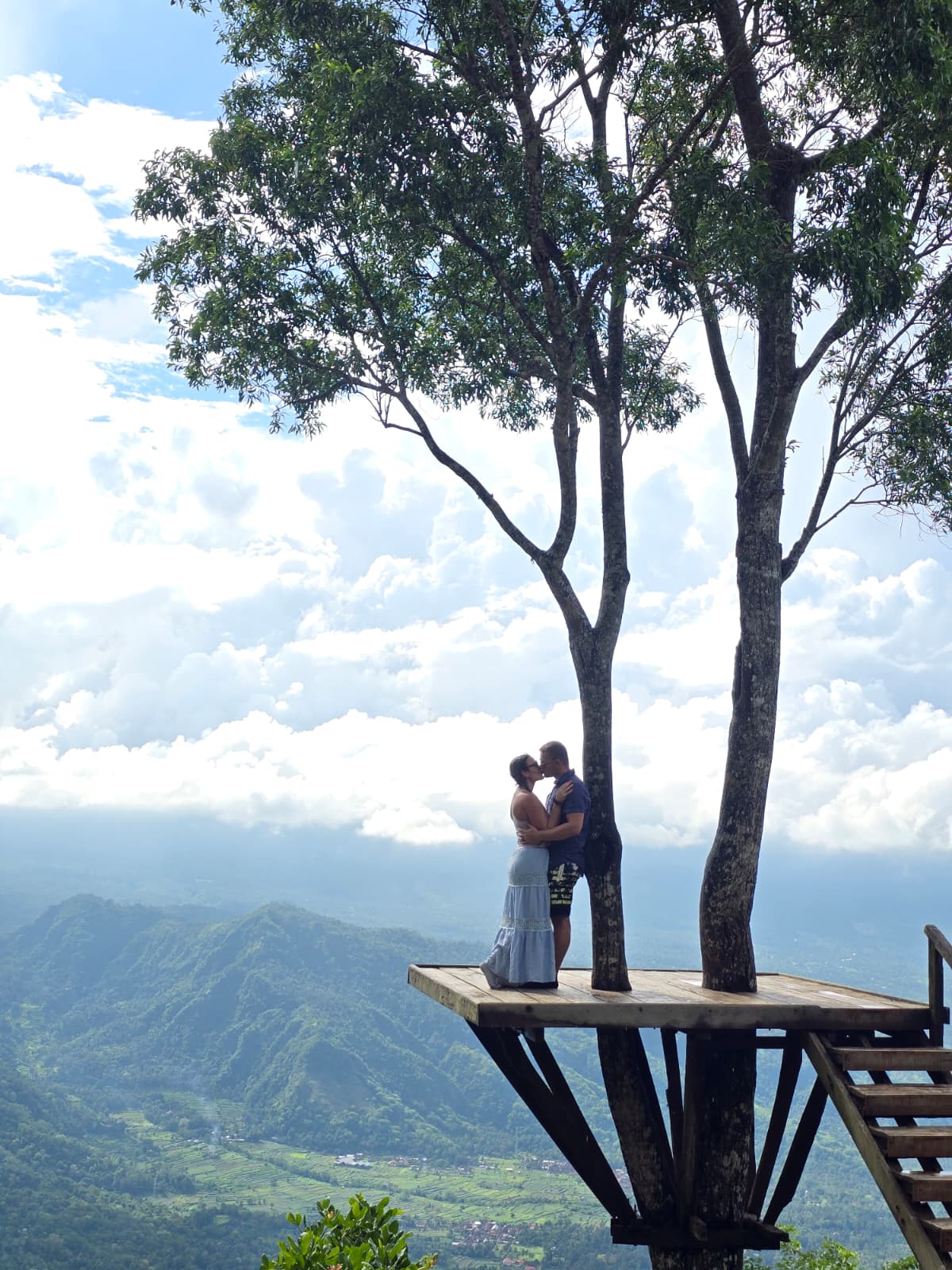Two people embrace on a wooden platform under a large tree with a mountain landscape in the background.