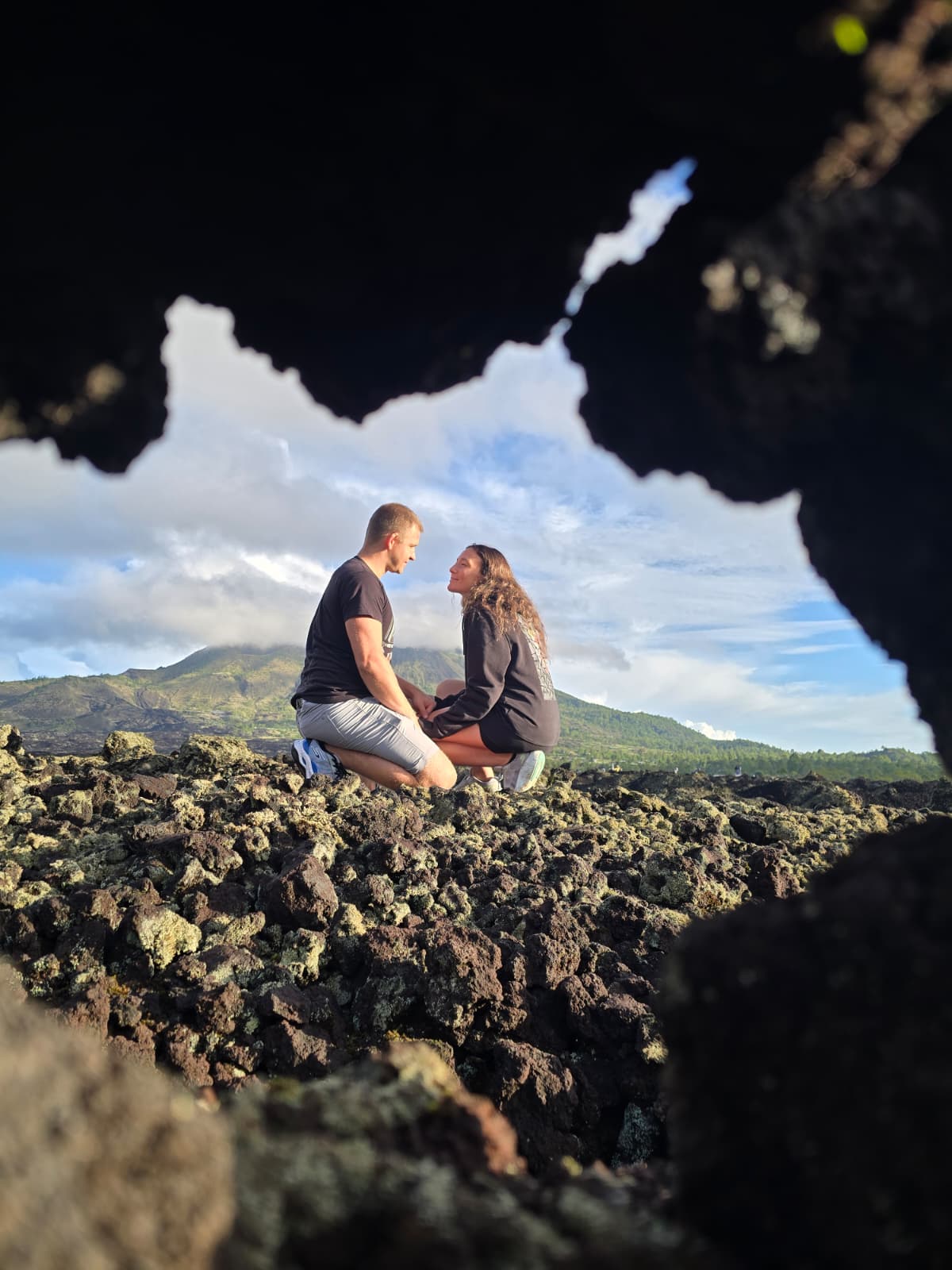 A couple kneeling on rocky terrain, holding hands, with mountains and sky in the background.