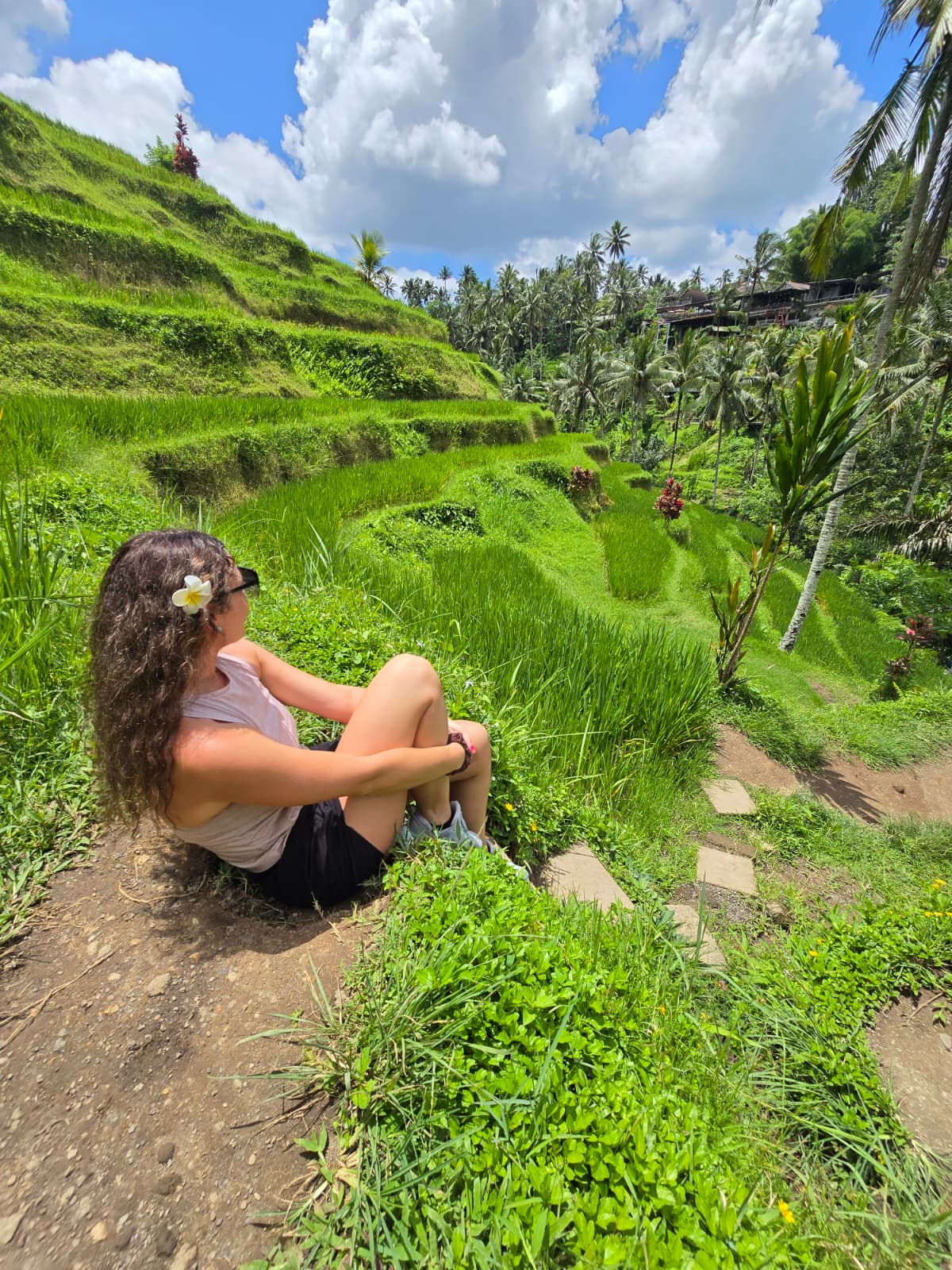 Girl with long hair and a flower in her hair sits on a dirt path in a lush green terraced landscape under a partly cloudy sky.