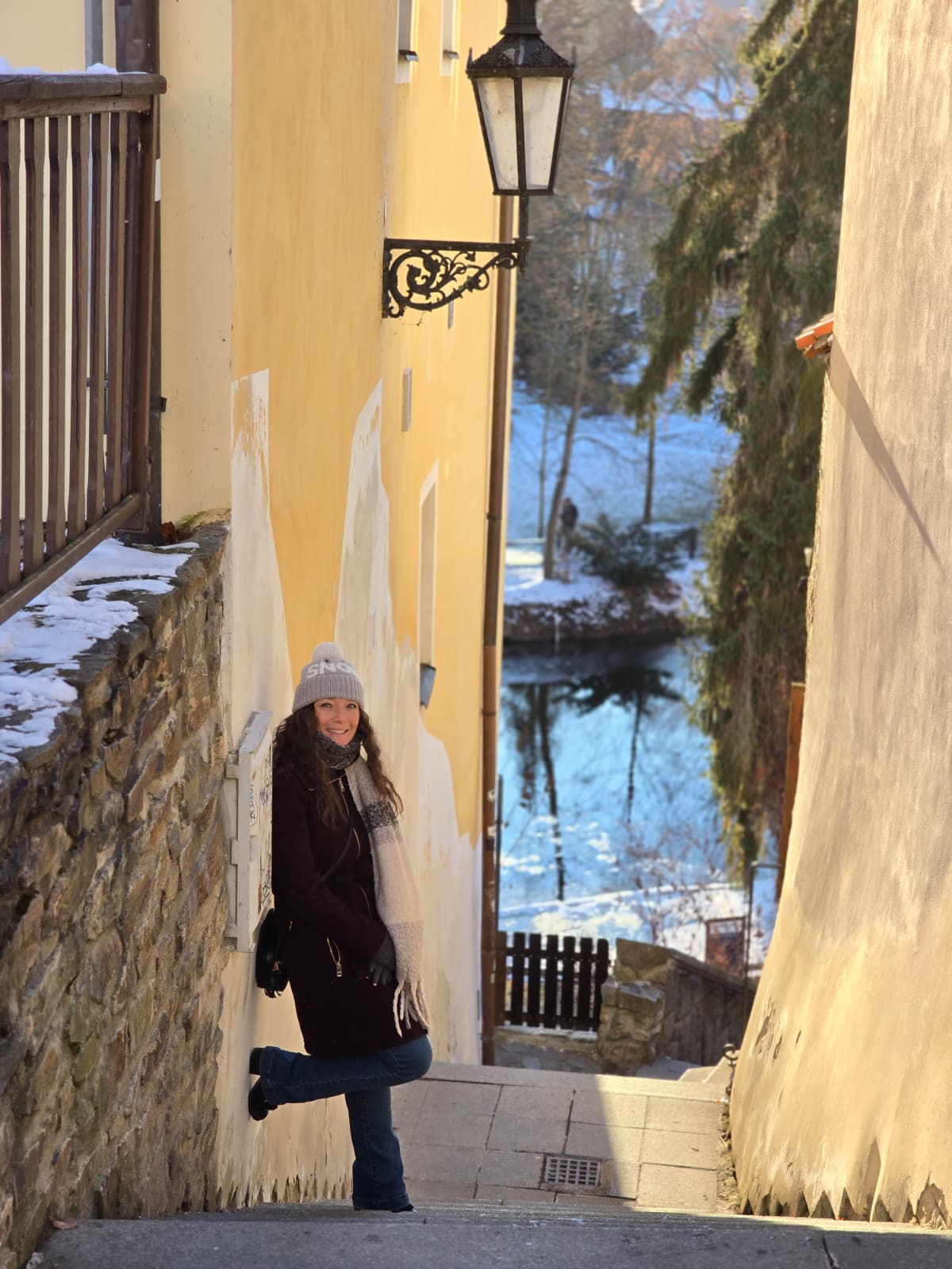 A woman wearing a gray hat and black coat stands on a narrow street with yellow and stone walls, snow, and a river in the background.