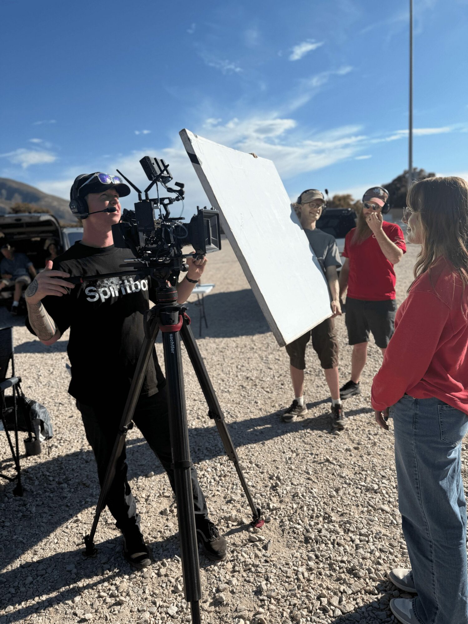 Group of people outdoors with camera crew, one person operating camera, others talking, bright sunny day, blue sky.