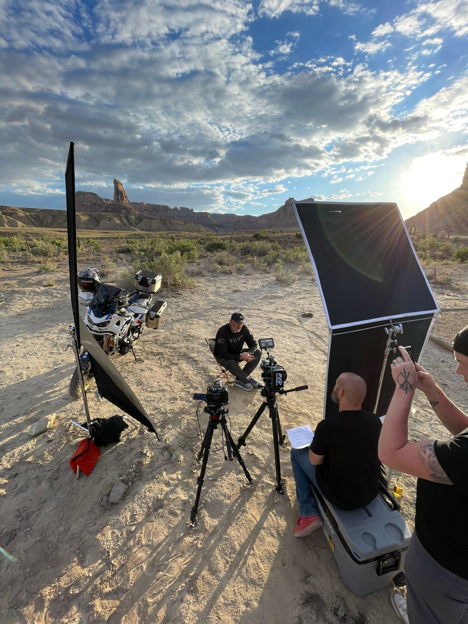 People setting up camera equipment outdoors on sandy terrain with a cloudy sky and distant hills.