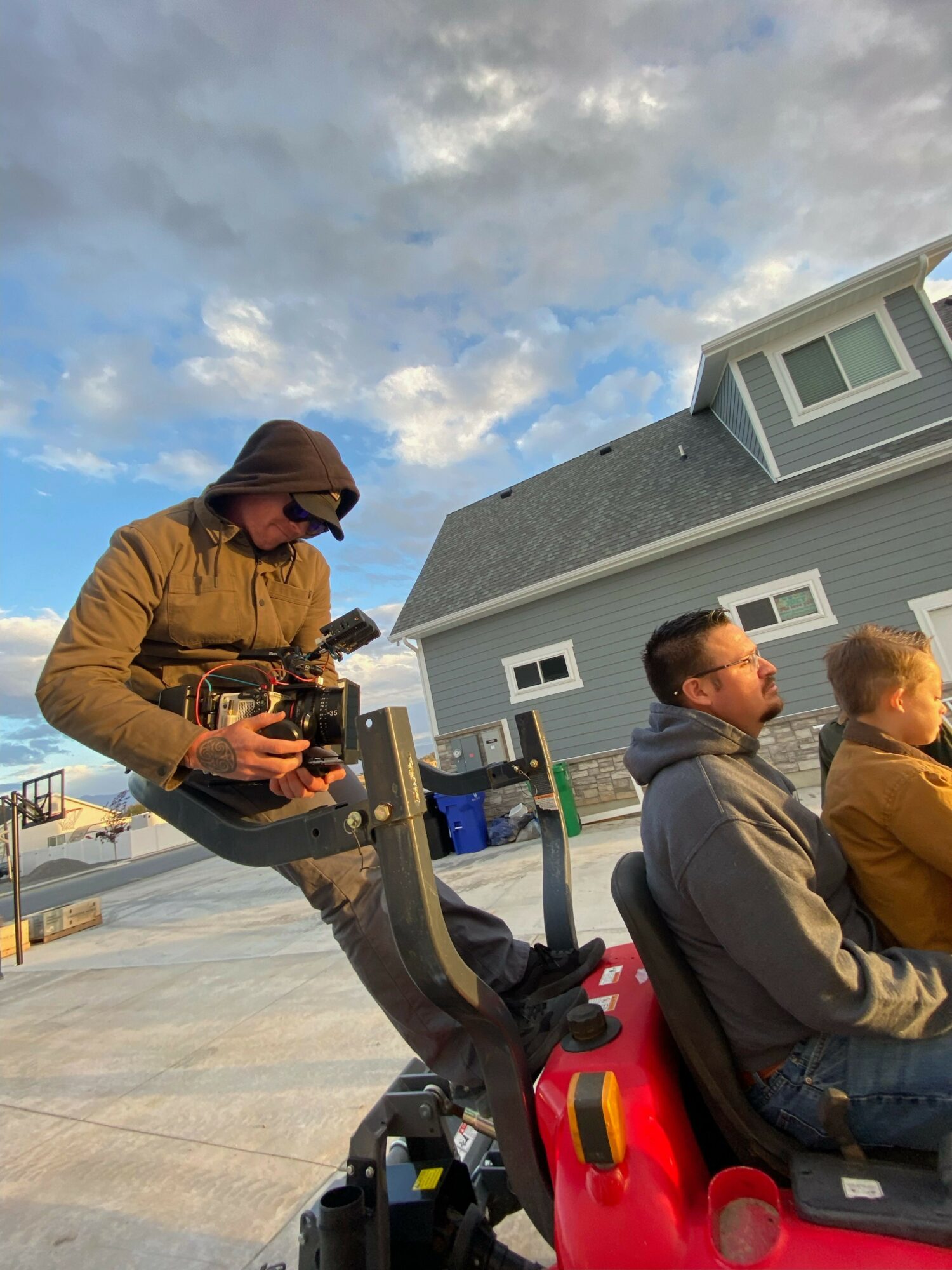 Three people sitting outdoors on a sunny day, one holding a camera, with a building and blue sky in background.