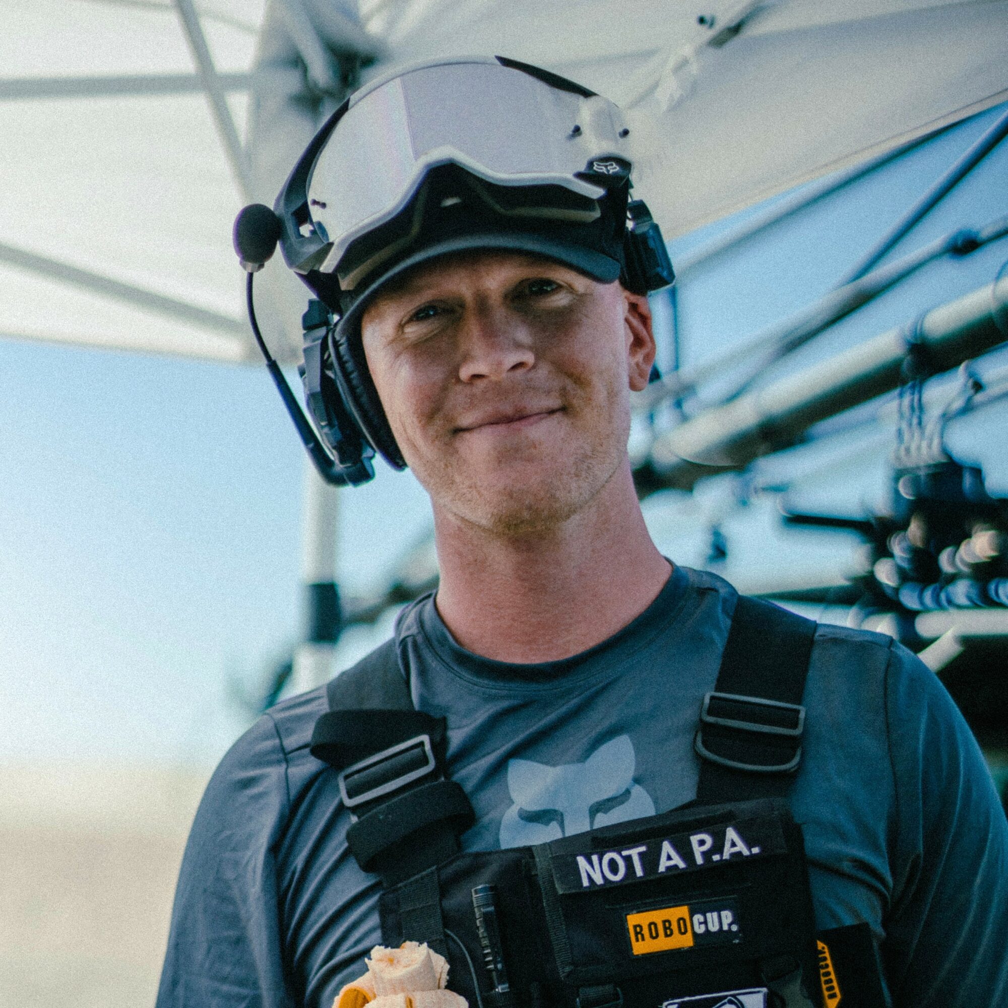 Young man wearing a helmet and headset, smiling, outdoors with a boat in the background.