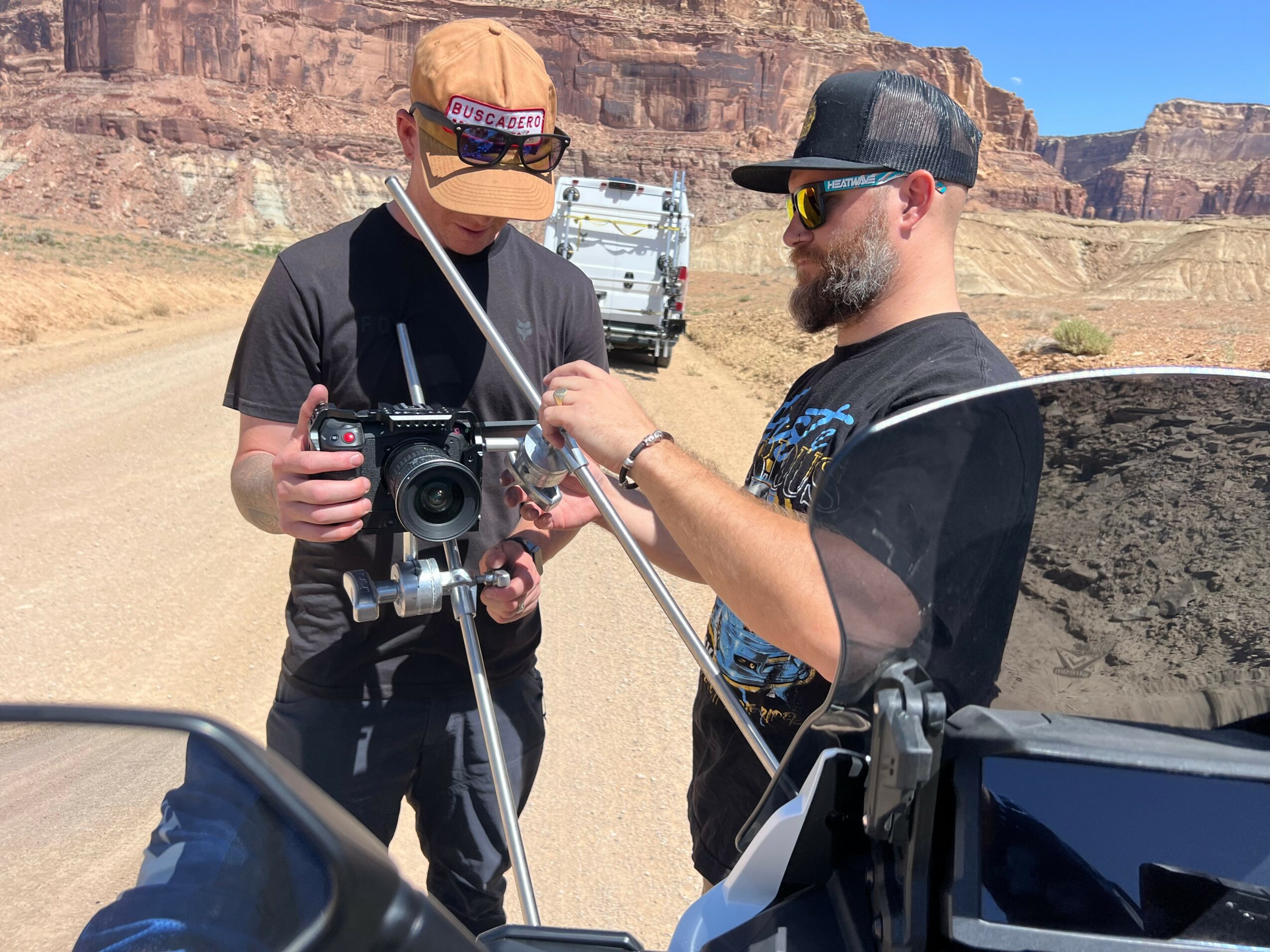 Two men stand outdoors in a desert landscape, working with a drone controller and equipment, with a vehicle and canyon in background.