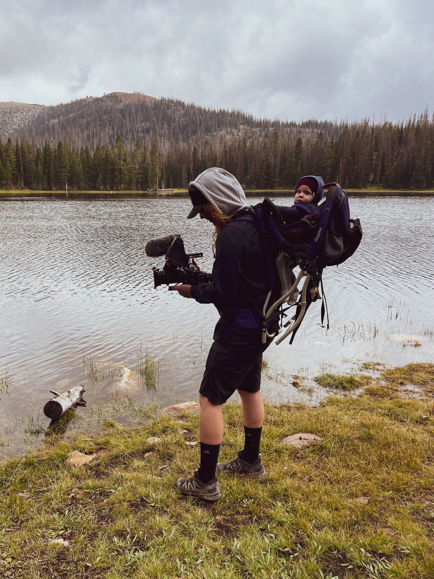 Person with a backpack and child in a carrier, holding a camera near a lake with trees and mountains in background