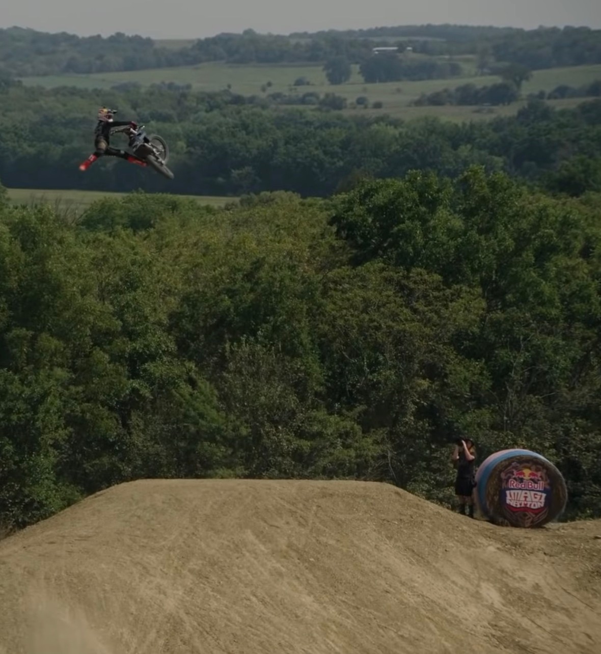 Motorcycle airborne above dirt jump with person standing nearby and trees in background.