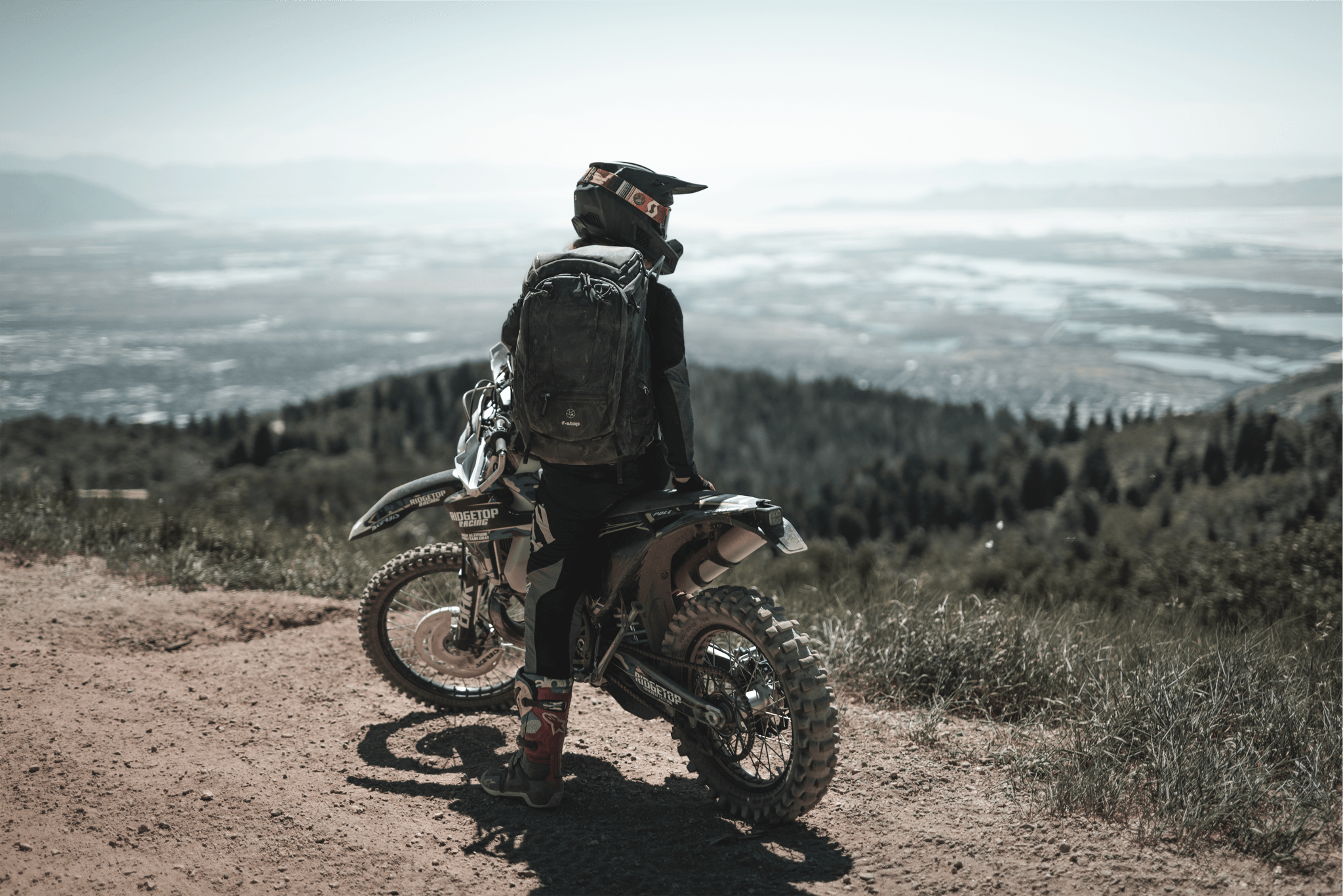 Person in motorcycle gear standing next to a dirt bike on a mountain trail, overlooking a landscape with water and forests.