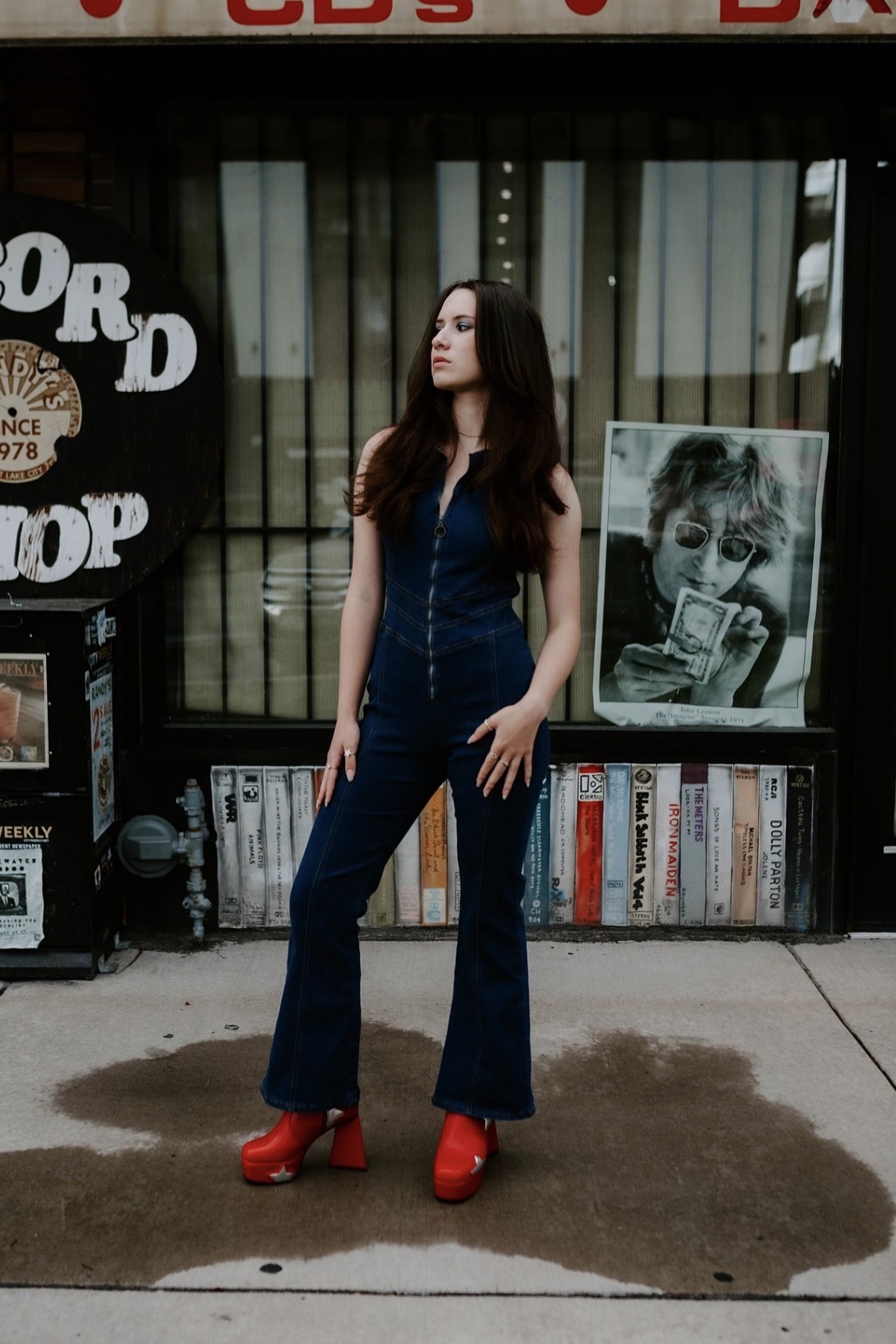 Woman with long dark hair in a dark jumpsuit and red shoes standing outside a bookstore, with a poster and books behind her.