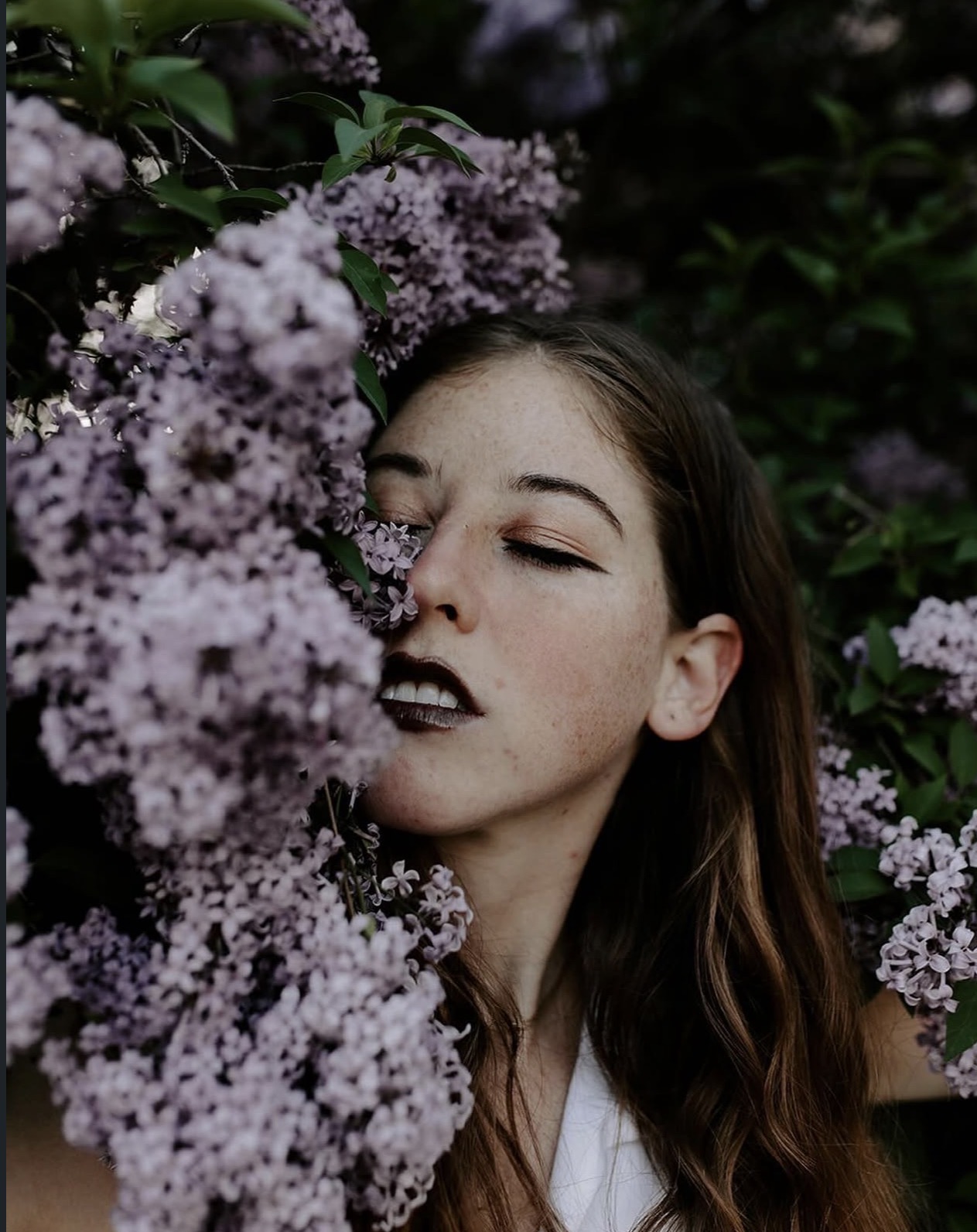 Young woman with long brown hair and dark lipstick among purple flowers, eyes closed, serene expression.