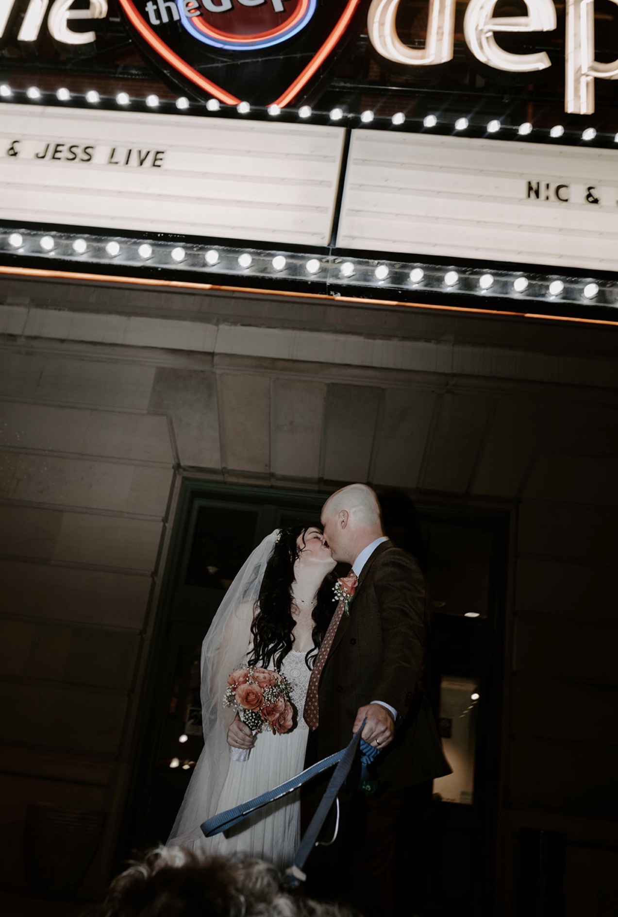 A bride and groom kiss outside a building at night, with the bride holding a bouquet and the groom holding a blue umbrella.