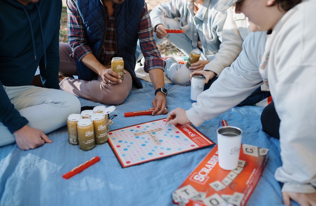 Group of people playing a board game on a blue tablecloth, holding drinks, with game pieces and snacks visible.
