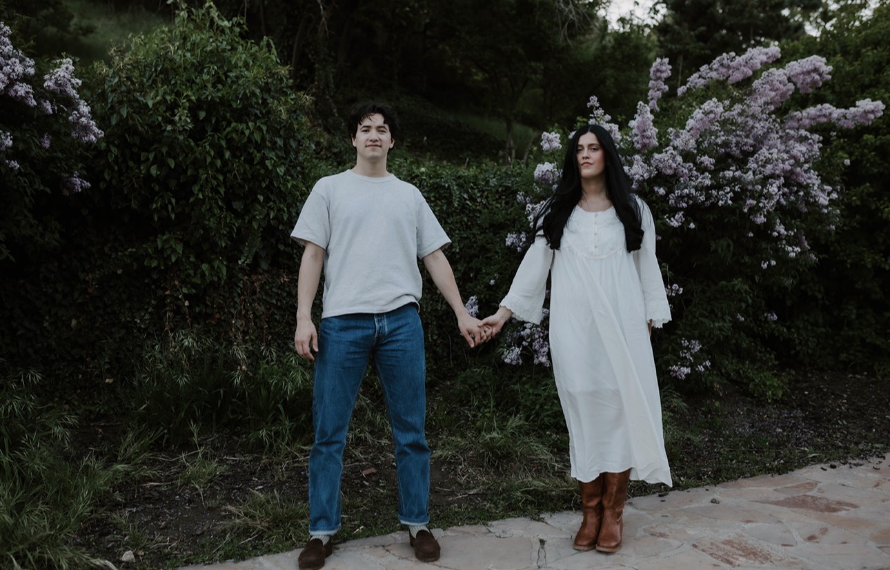 A young man and woman holding hands outdoors near flowering bushes, standing on a sidewalk with trees in the background.