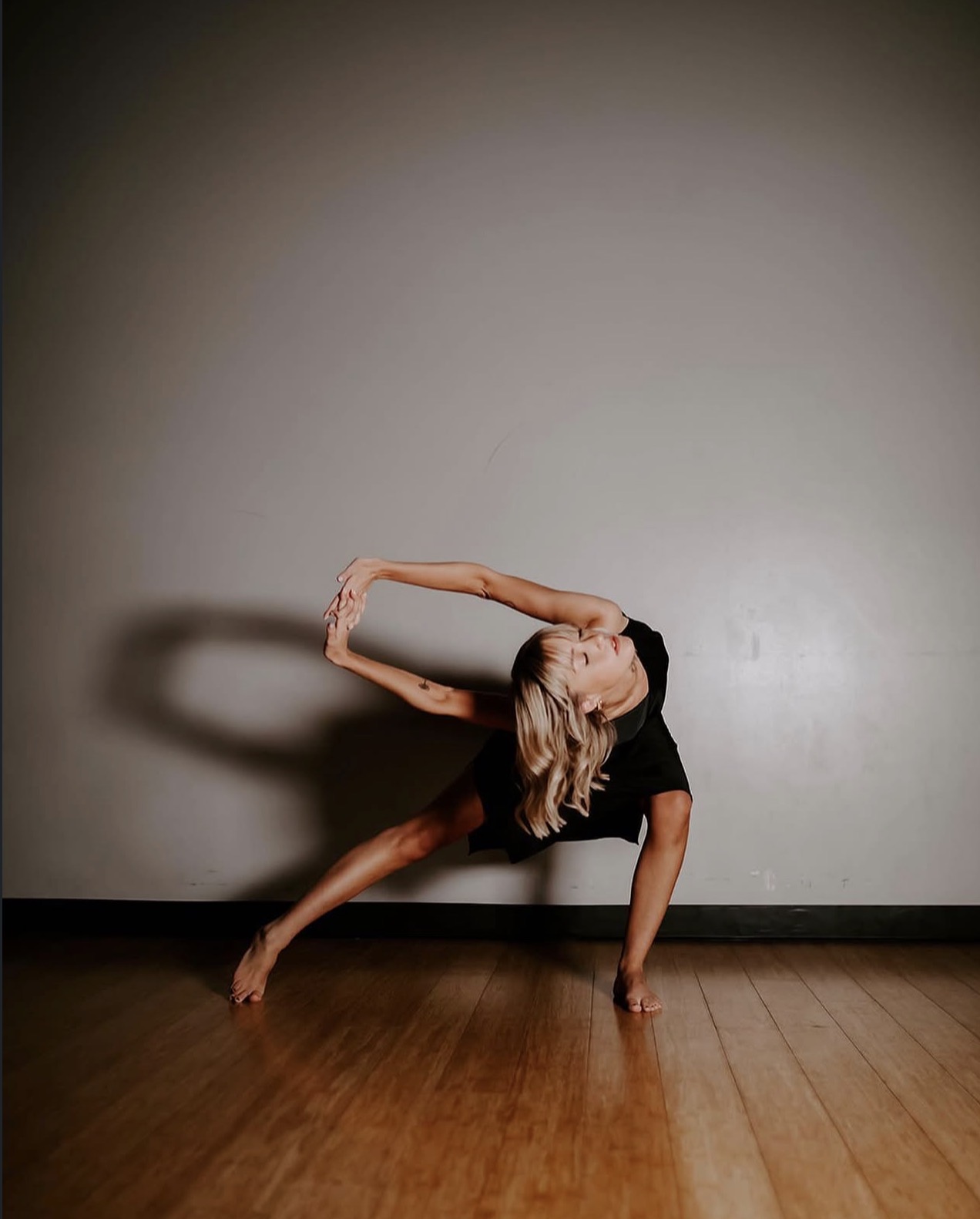 Person performing a side bend stretch with one arm overhead, balancing on one hand and foot, in a dance studio.