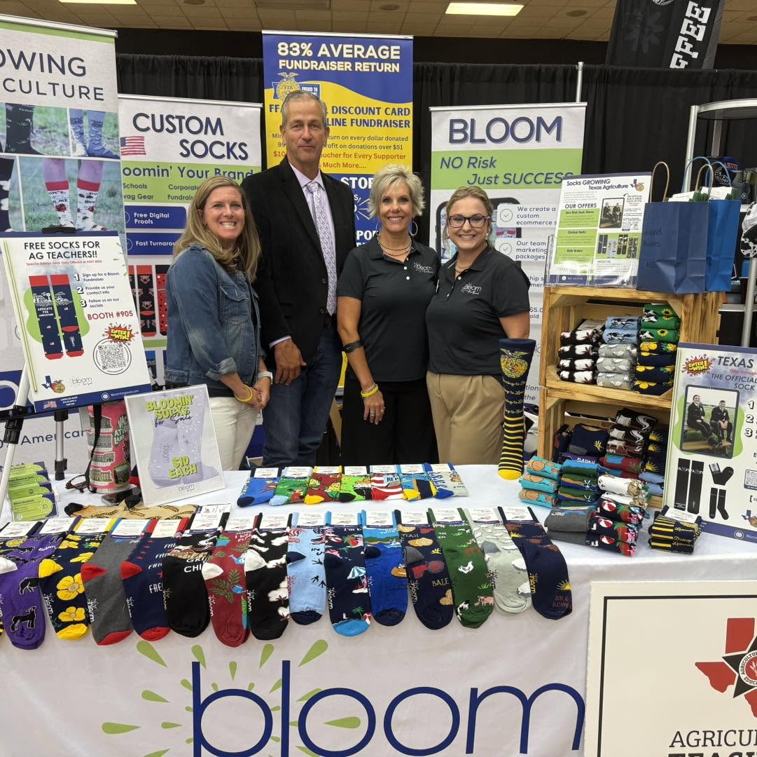 Four people stand behind a table displaying colorful socks at a trade show booth, with banners and product displays around them.
