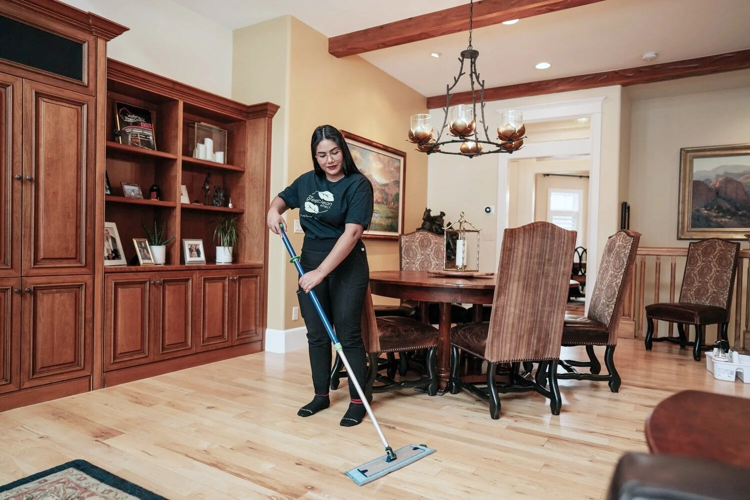 Woman mopping wooden floor in dining room with large table and chairs, wooden shelves, and chandelier, in a well-lit home interior.