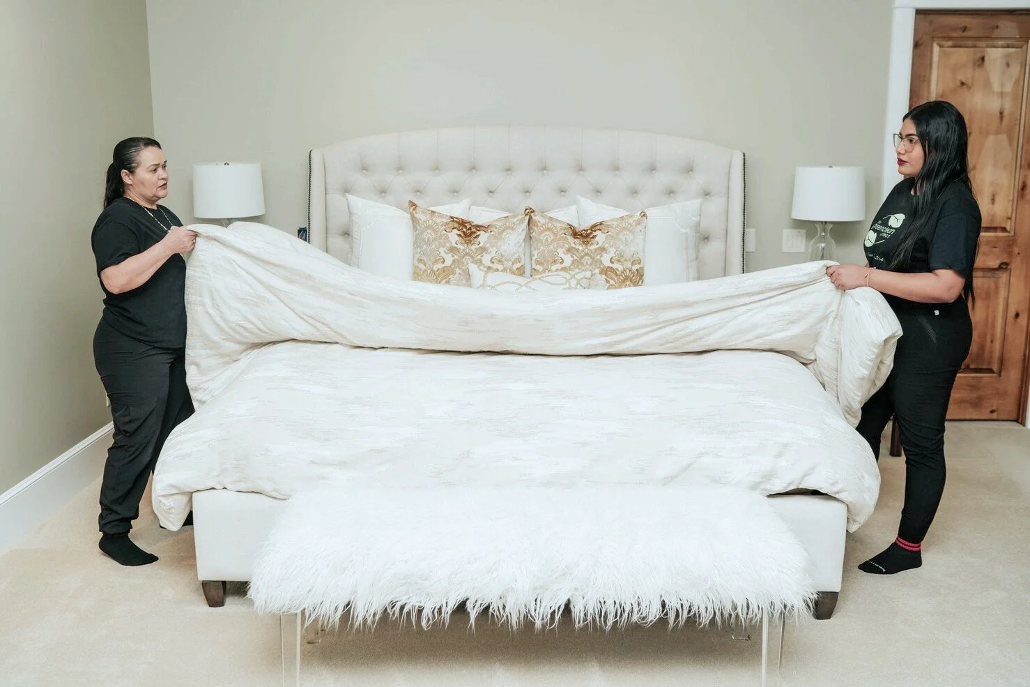 Two women make a bed with white bedding in a bedroom with green walls and wooden furniture.