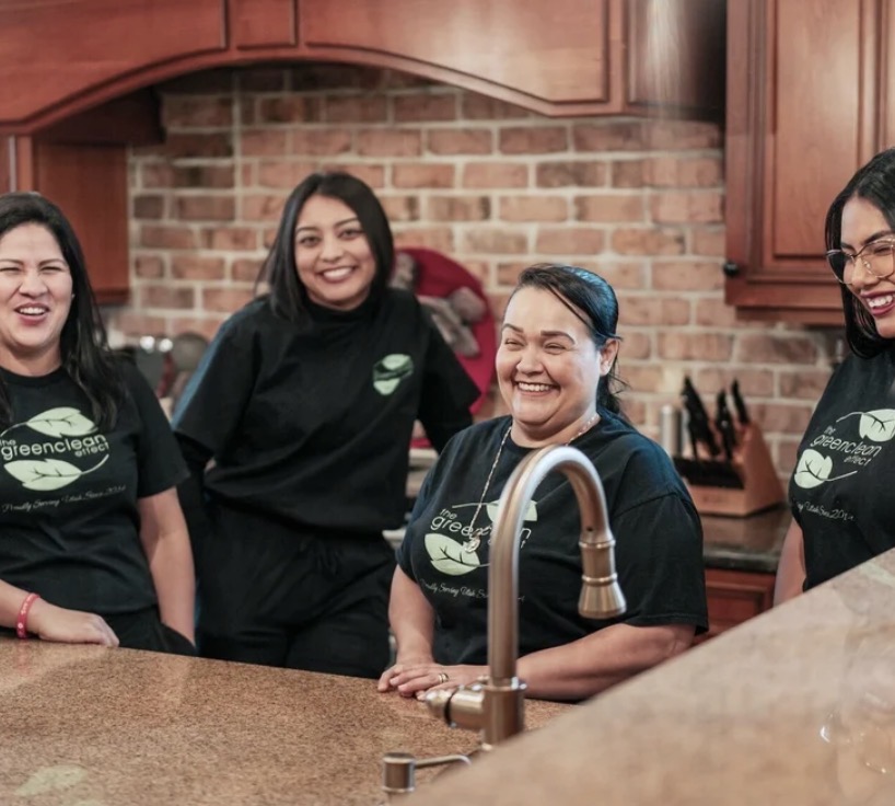 Four women smiling in a kitchen with brick wall and wooden cabinets, wearing black shirts with green logos.