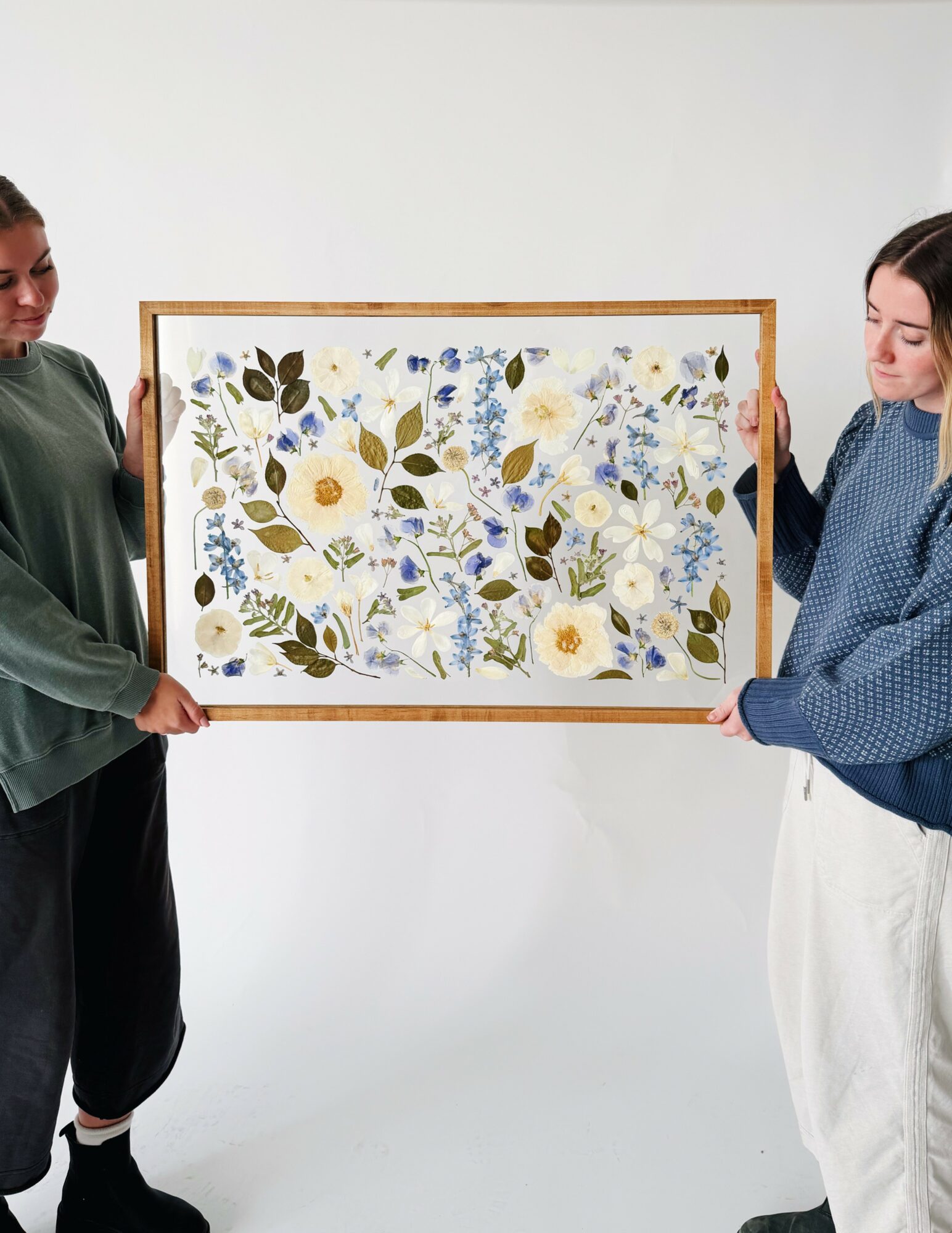Two women hold a framed floral artwork with blue, yellow, and green flowers against a white background.