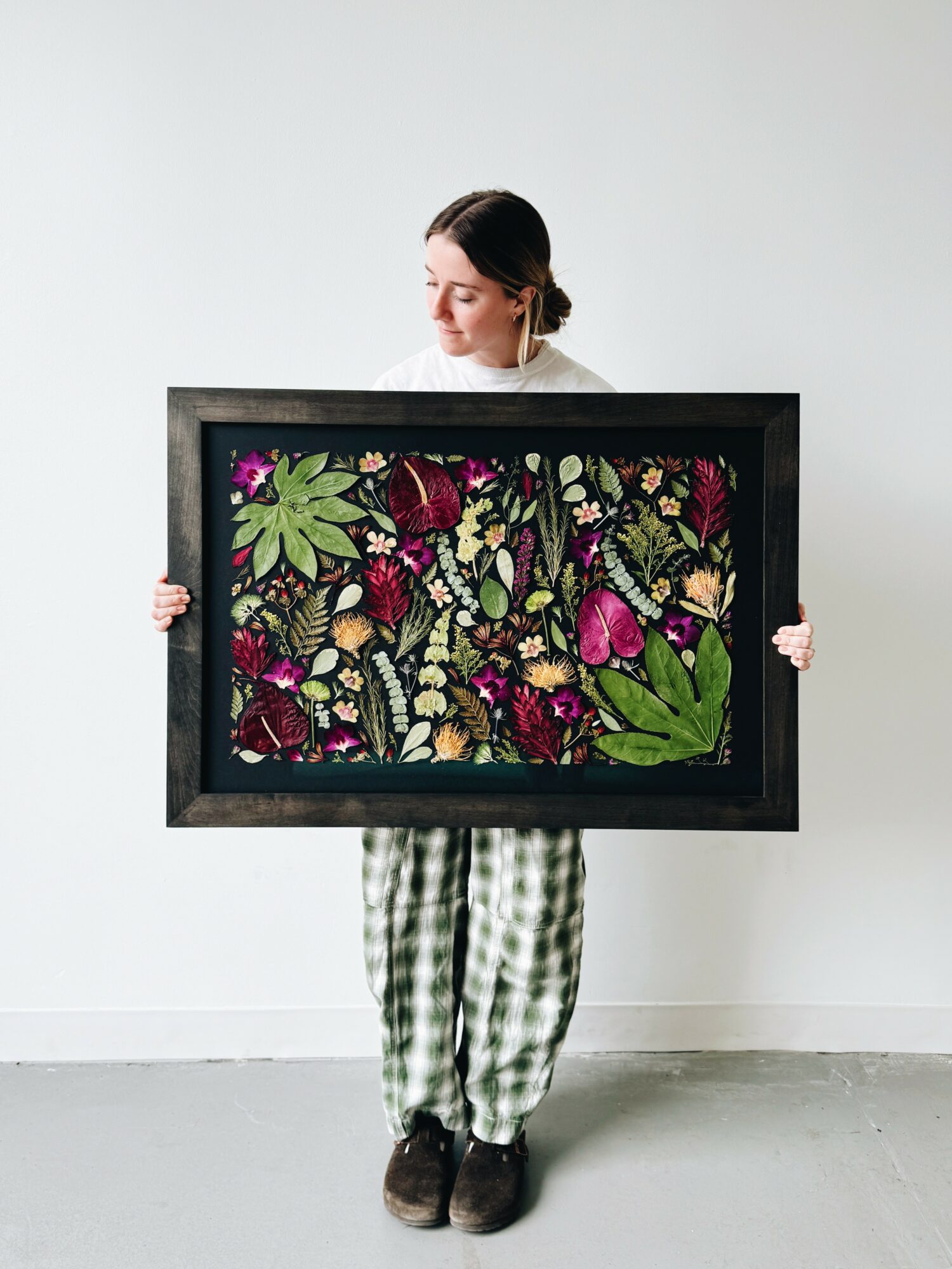 Person holding a framed colorful floral artwork, standing against a plain white wall, wearing checkered pants and dark shoes.