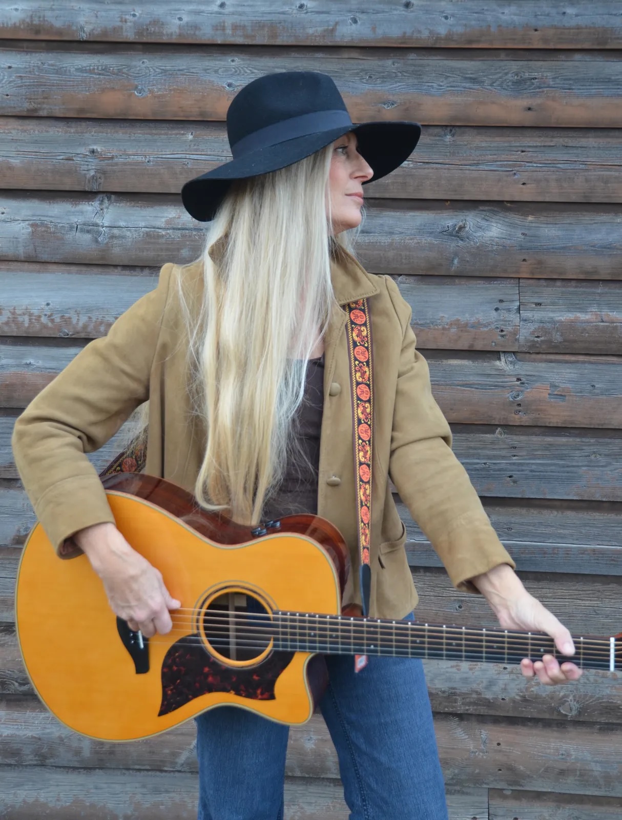 Woman with long blonde hair wearing a black hat and tan coat playing an acoustic guitar against a wooden wall.