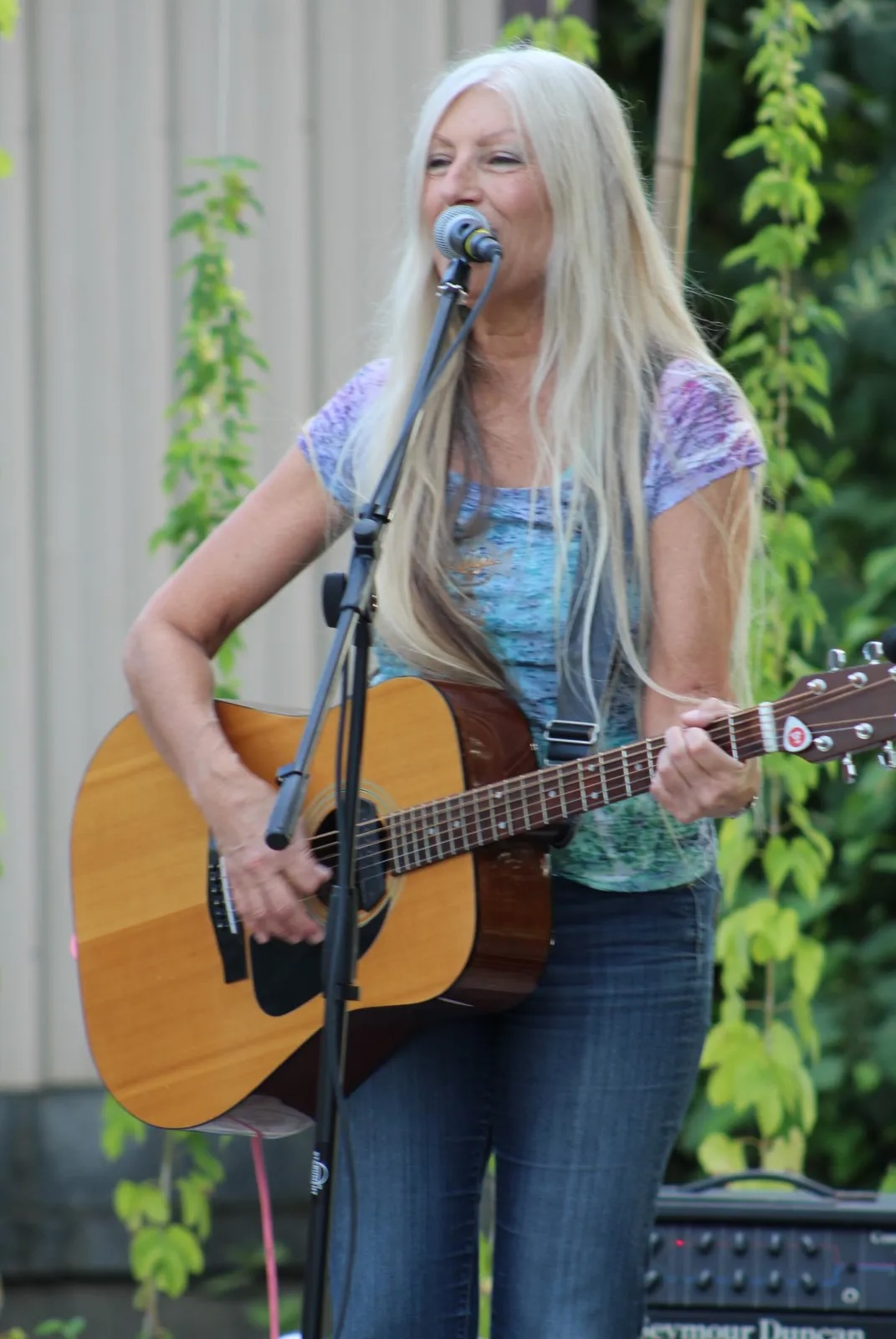 Woman with long blonde hair singing into a microphone while playing an acoustic guitar outdoors, surrounded by green plants.