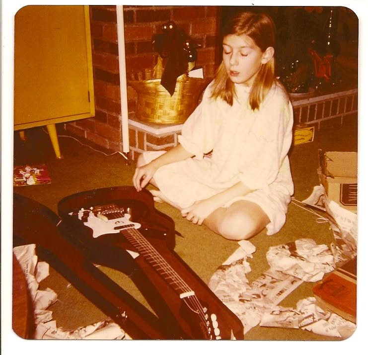 Girl sitting on the floor with a guitar case, surrounded by papers and objects, in a room with brick wall and yellow cabinet.
