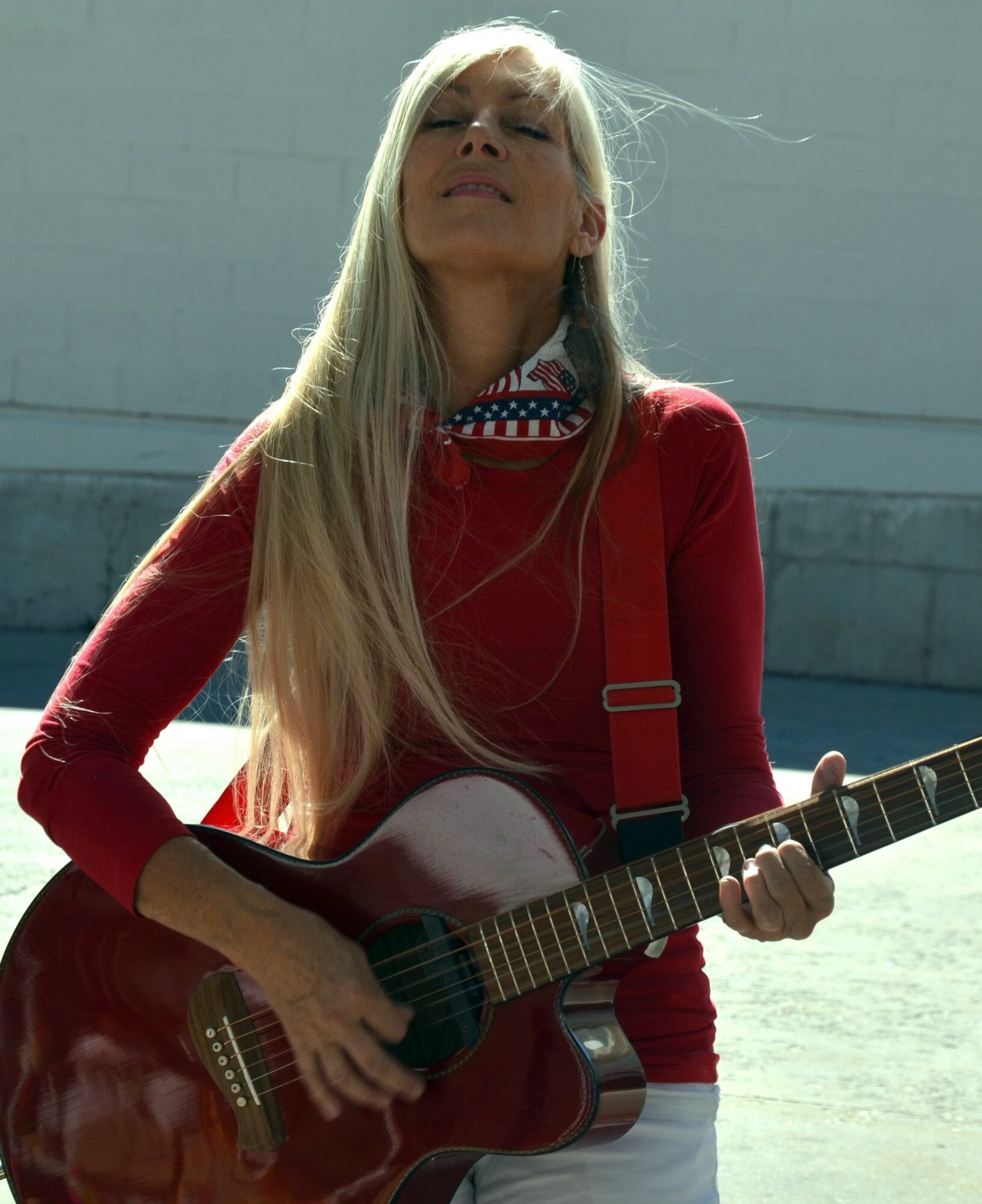 Woman with long blonde hair playing an acoustic guitar outdoors, wearing a red top and patterned scarf, sunlight on face.