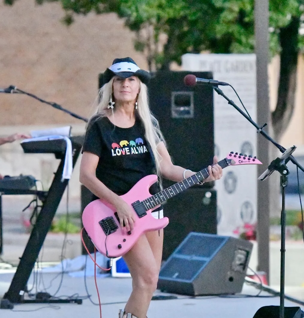 Young woman playing pink electric guitar outdoors, wearing a black T-shirt with colorful text, sunglasses, and a hat, with musical equipment around her.