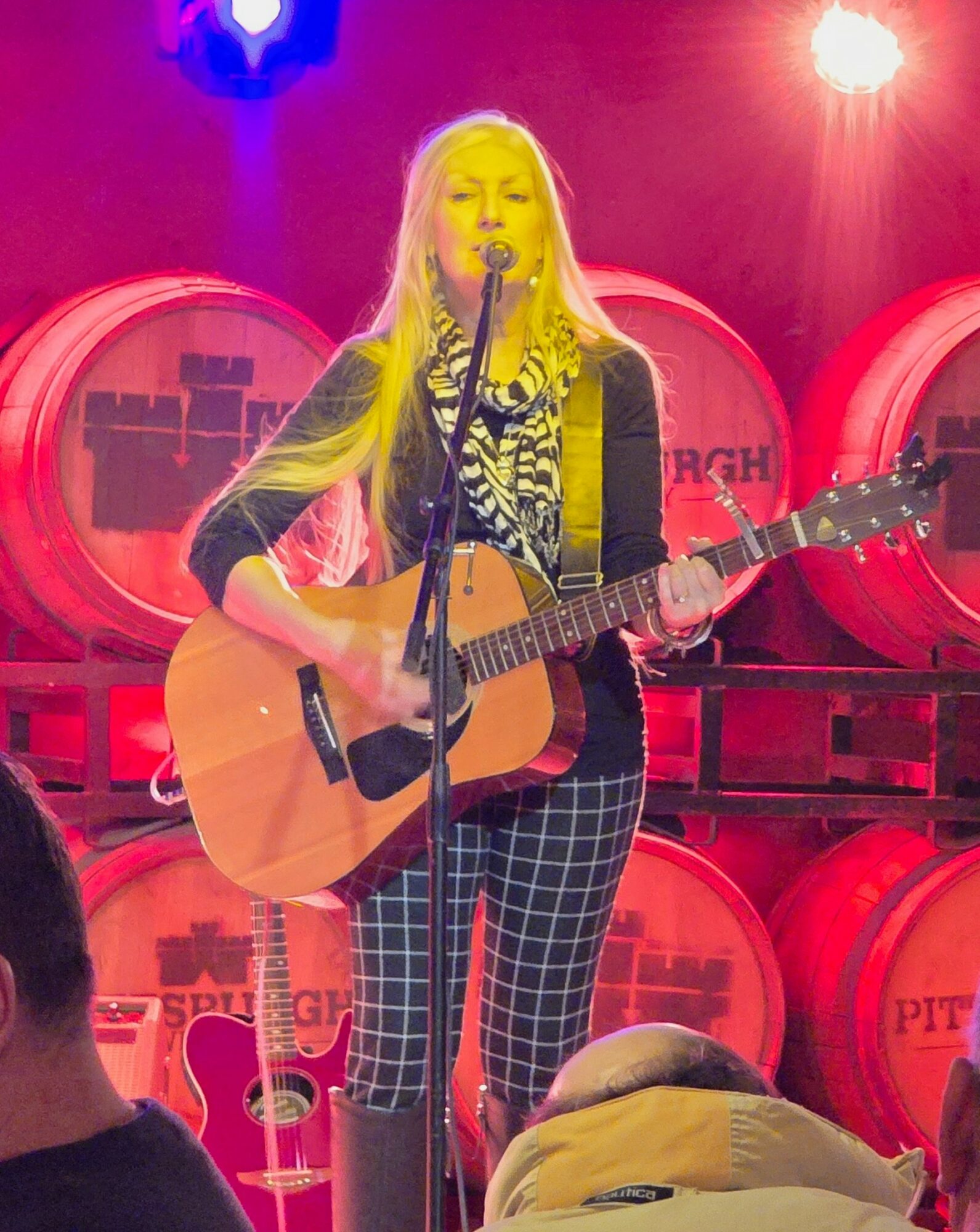 Woman with long blonde hair singing and playing guitar on stage with red barrels in background, pink and blue lighting.