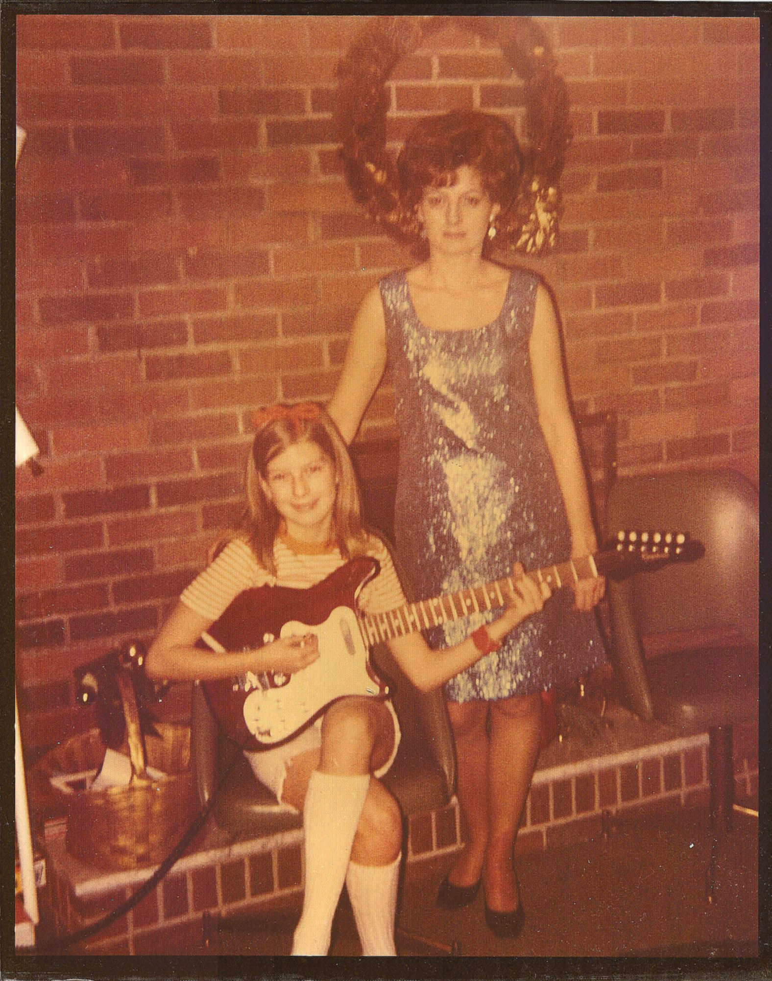 Two young girls, one sitting with an electric guitar and one standing, in front of a brick wall with a wreath.