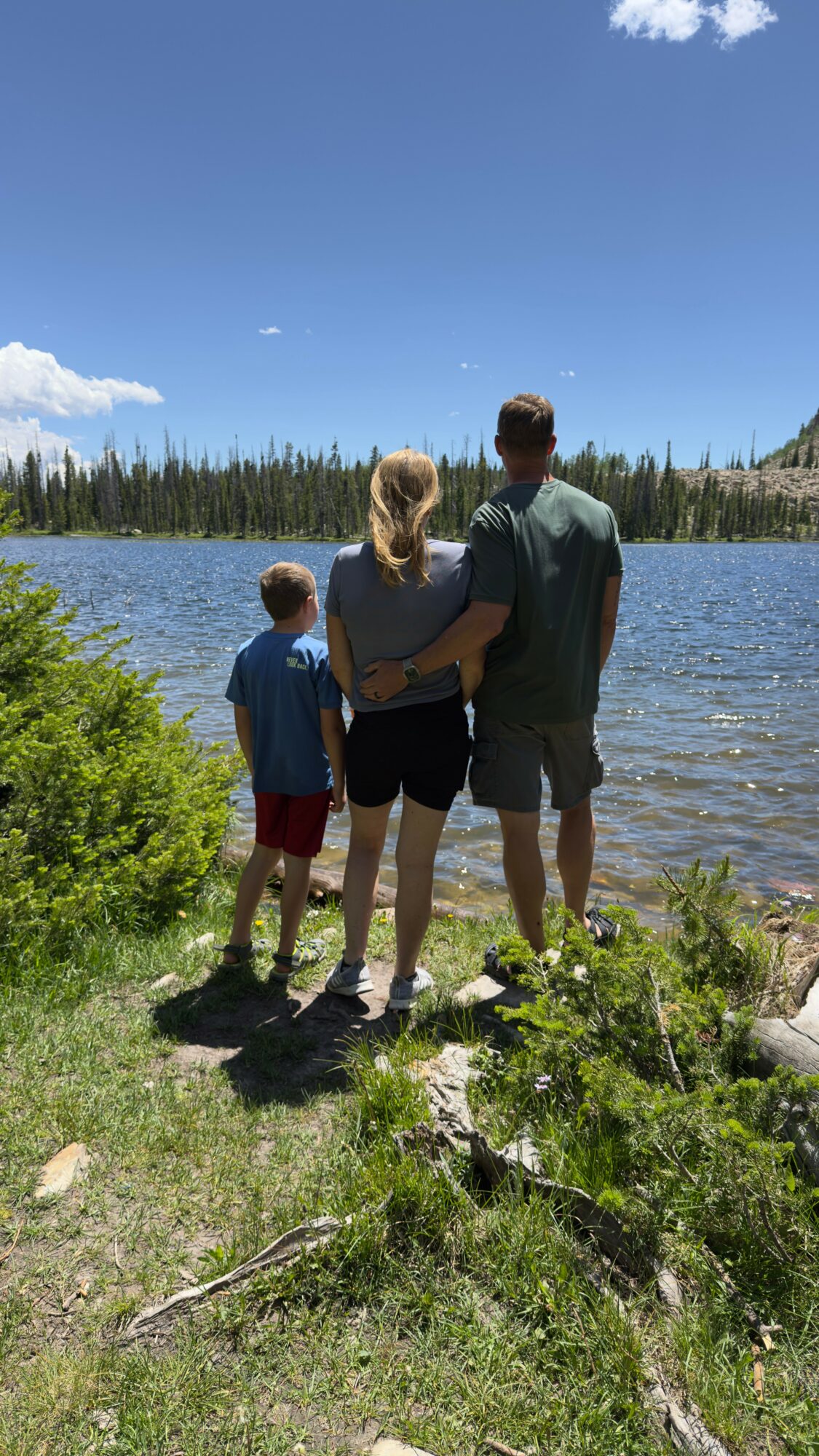 Family of three standing by a lake with trees and blue sky in the background, facing the water.