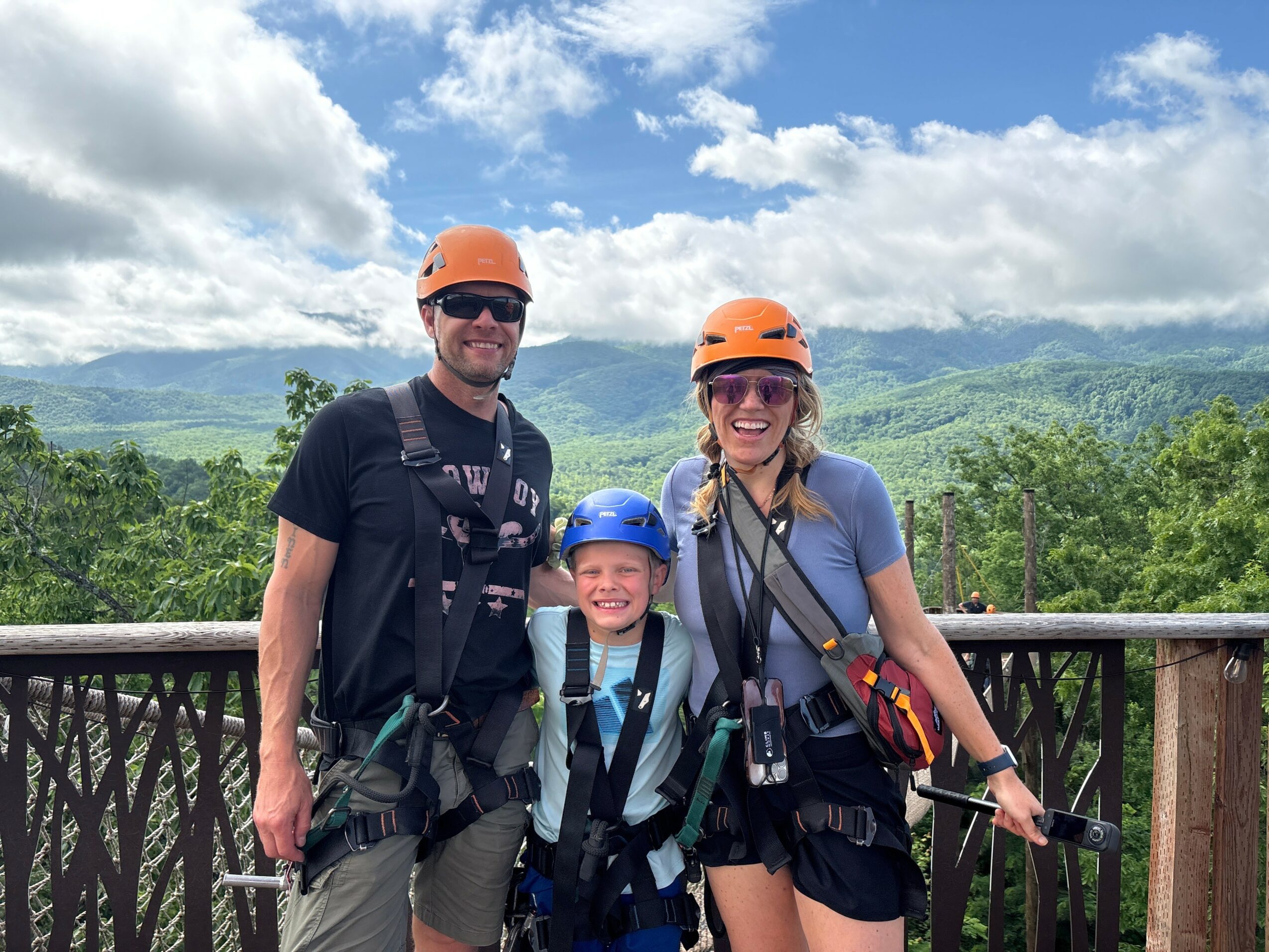 A family of three wearing helmets and harnesses outdoors with a scenic green landscape and cloudy sky behind them.
