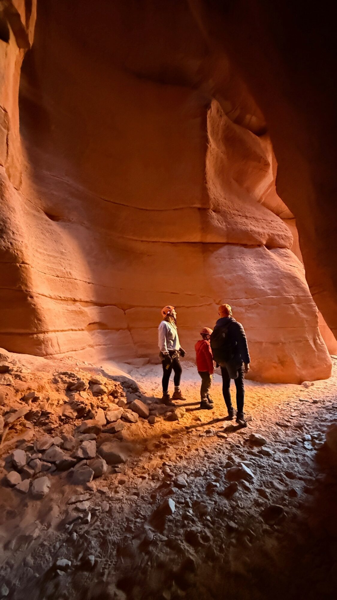 Three people stand inside a canyon with tall, smooth, reddish rock walls and a rocky ground.