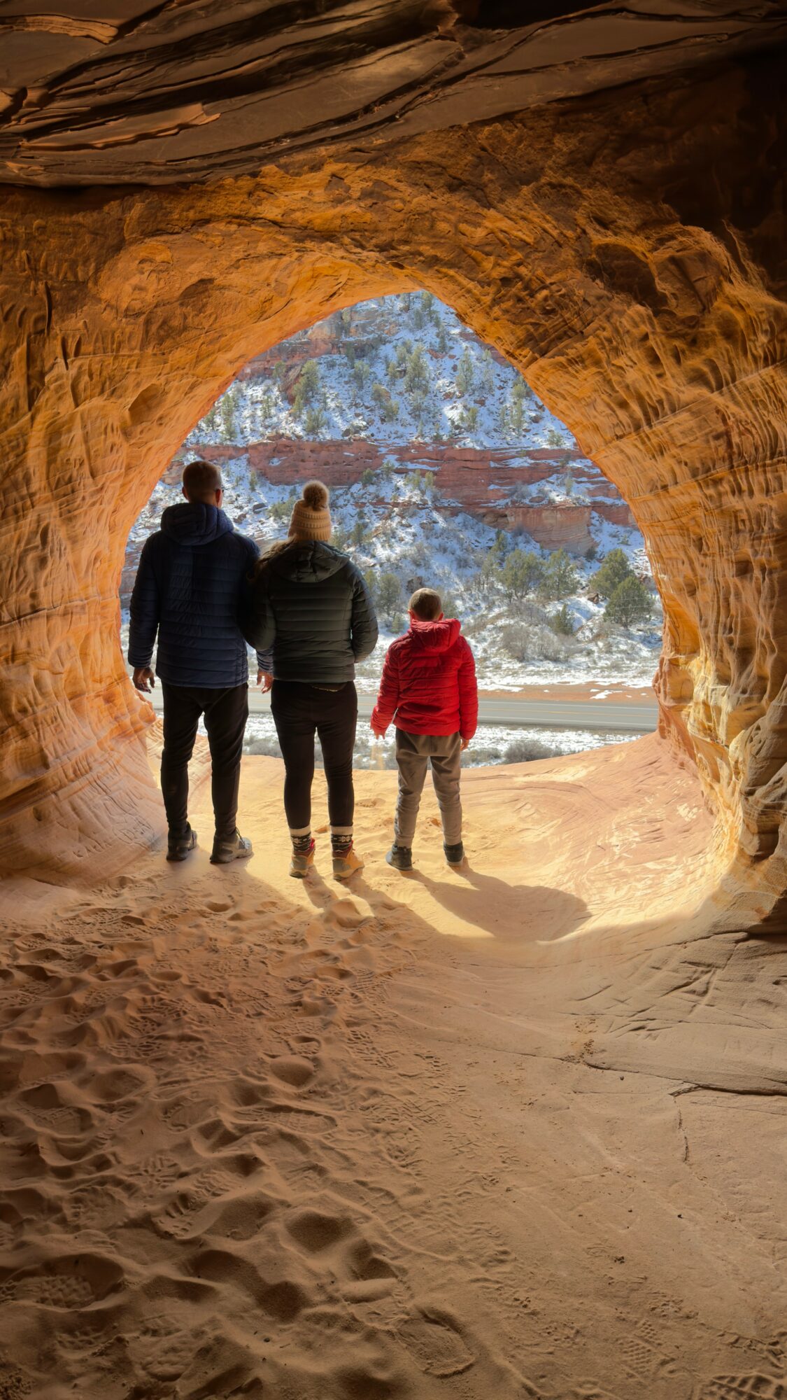 Three people, two adults and one child, walk through a cave opening toward a beach and ocean view.