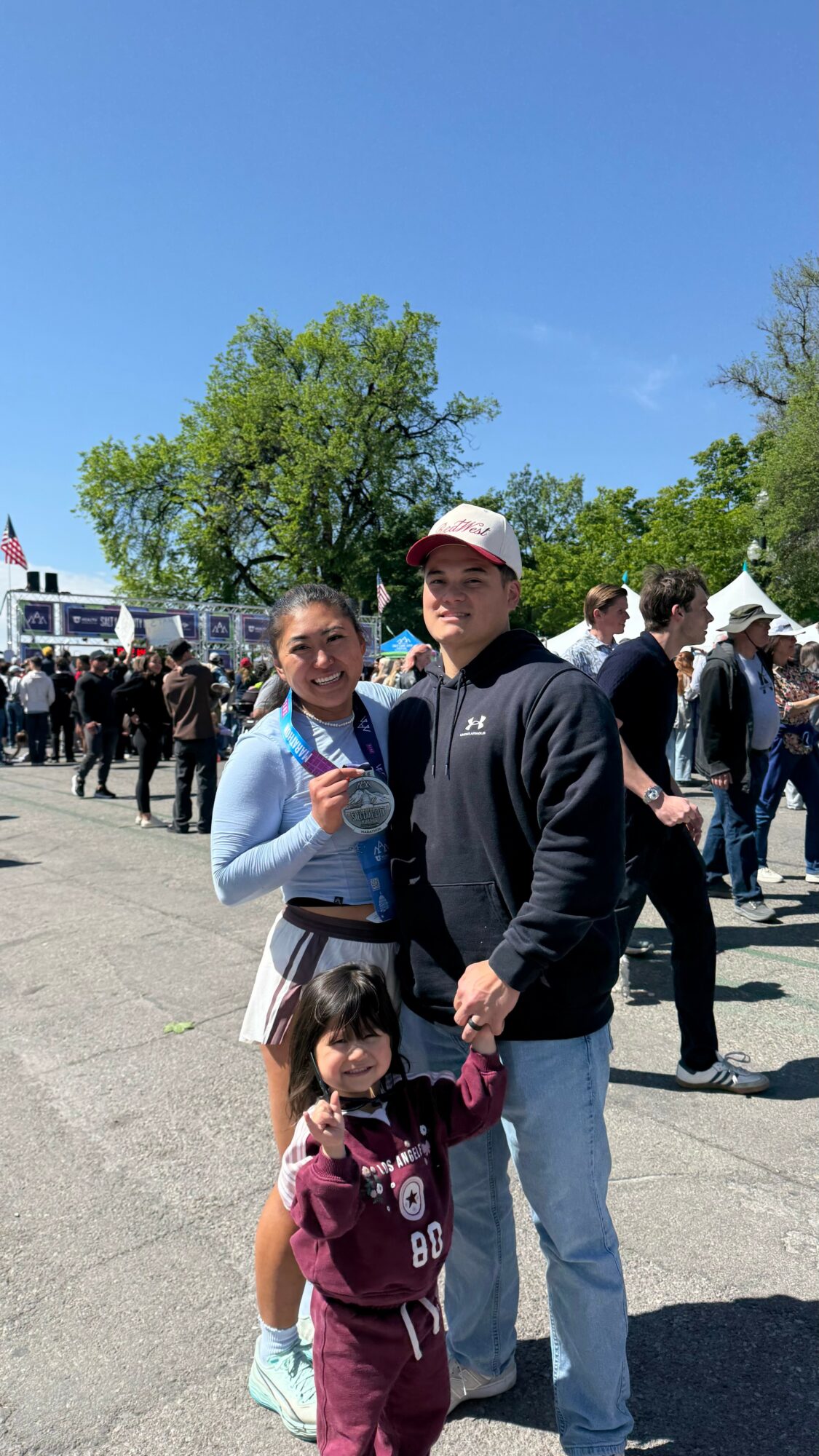 Three people, two adults and a child, standing outdoors with a crowd and trees in the background under a clear blue sky.