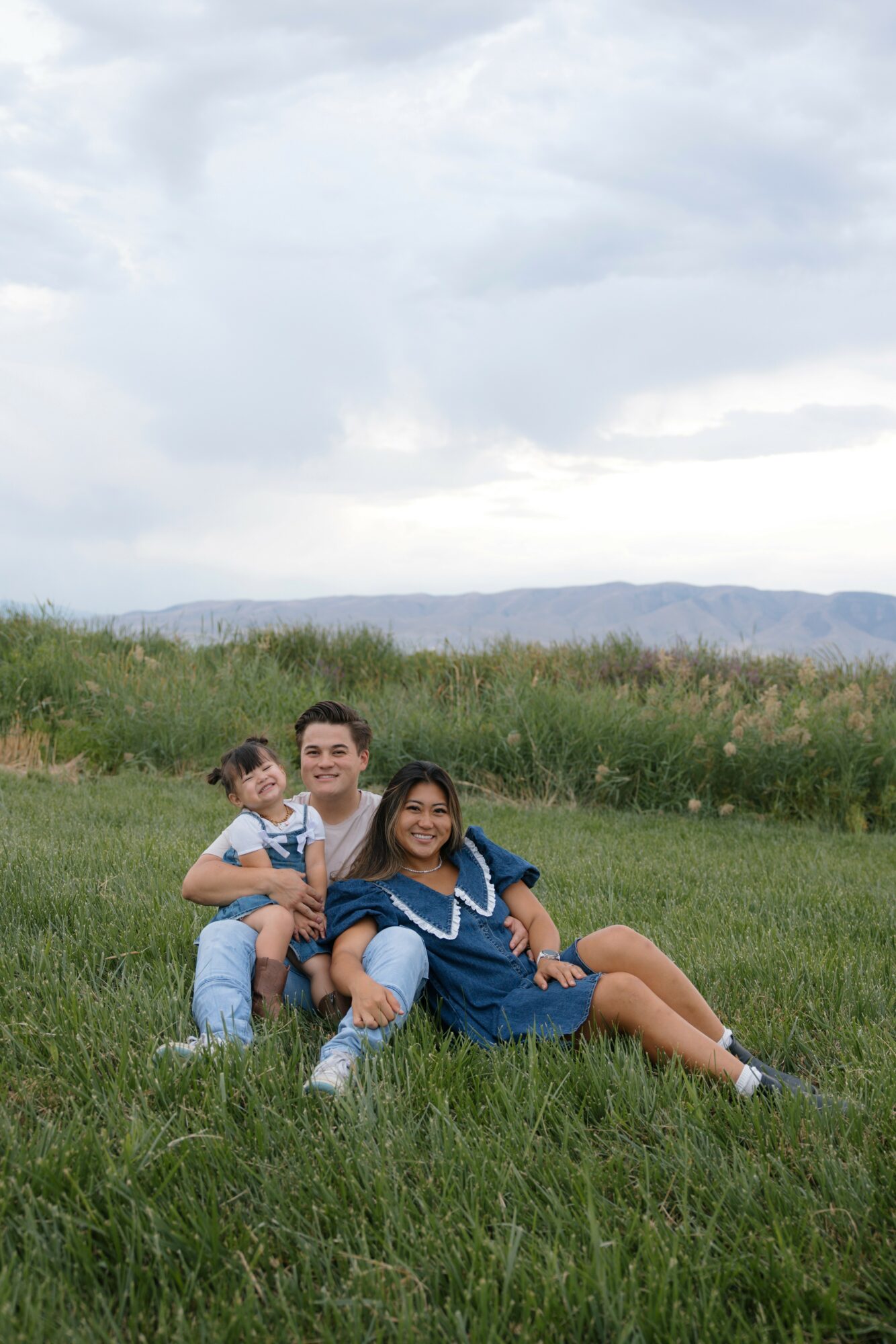 Three people sitting on grass in a field with hills and cloudy sky in background, smiling at camera.