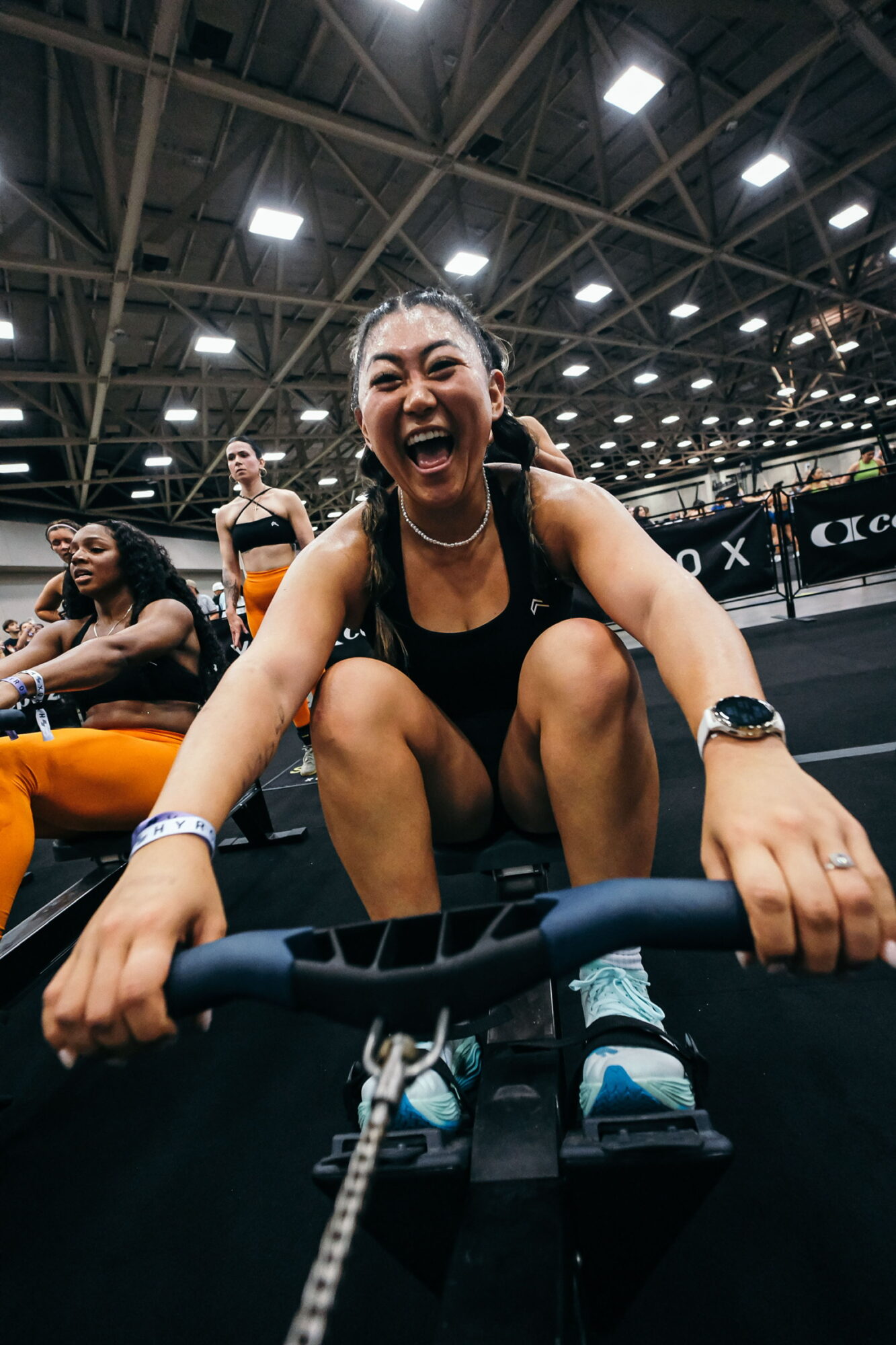 Woman smiling and laughing while riding a bike indoors, with other people and a high ceiling in the background.