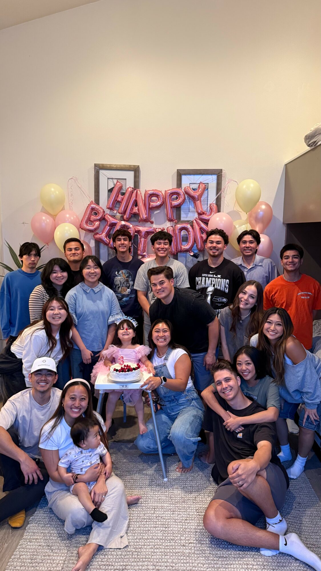 Group of people celebrating birthday with balloons and a cake, some children and adults smiling, in a room with a plain wall.