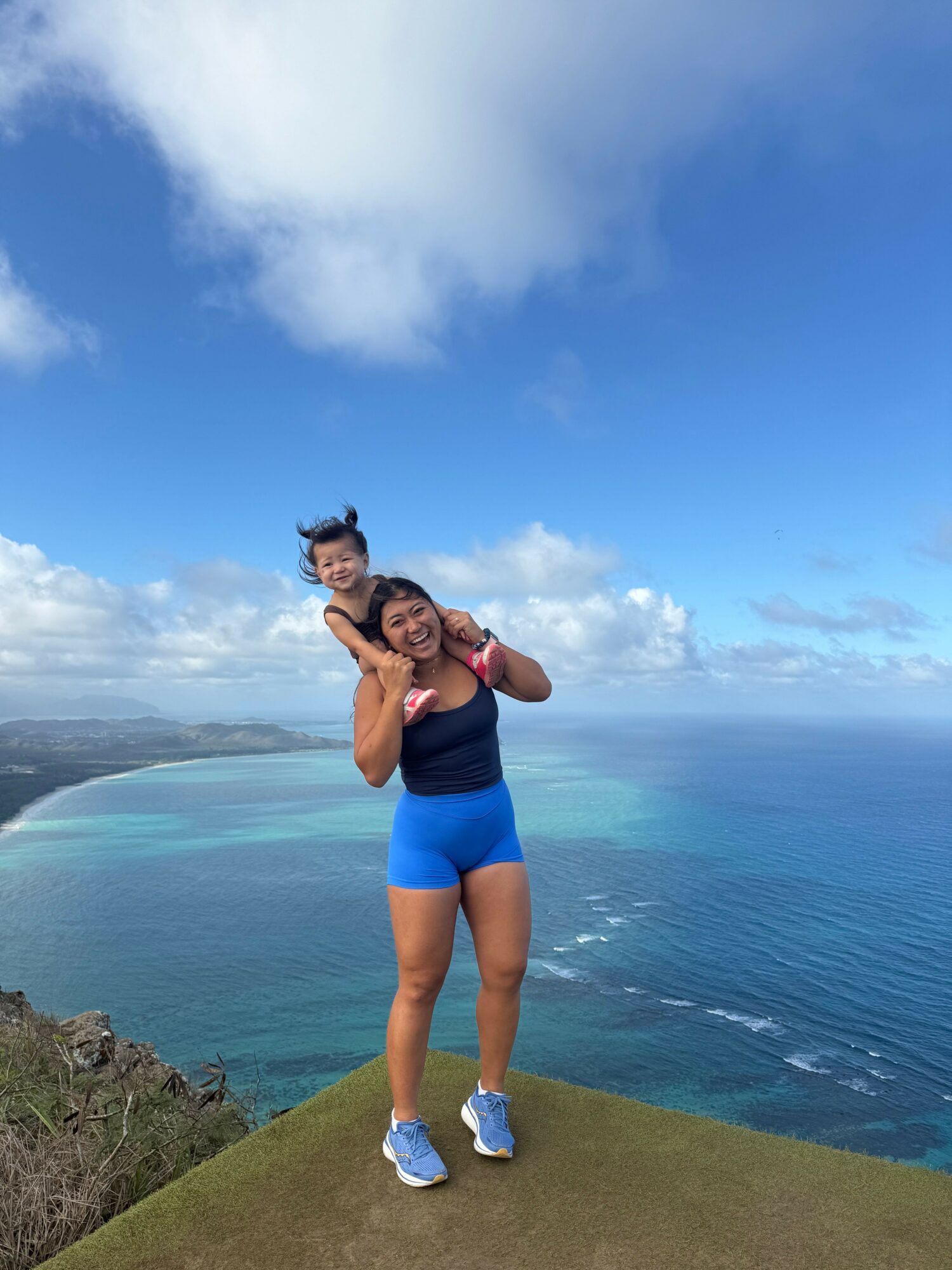 Woman standing on grass near ocean, holding a child, with blue sky and clouds in background.