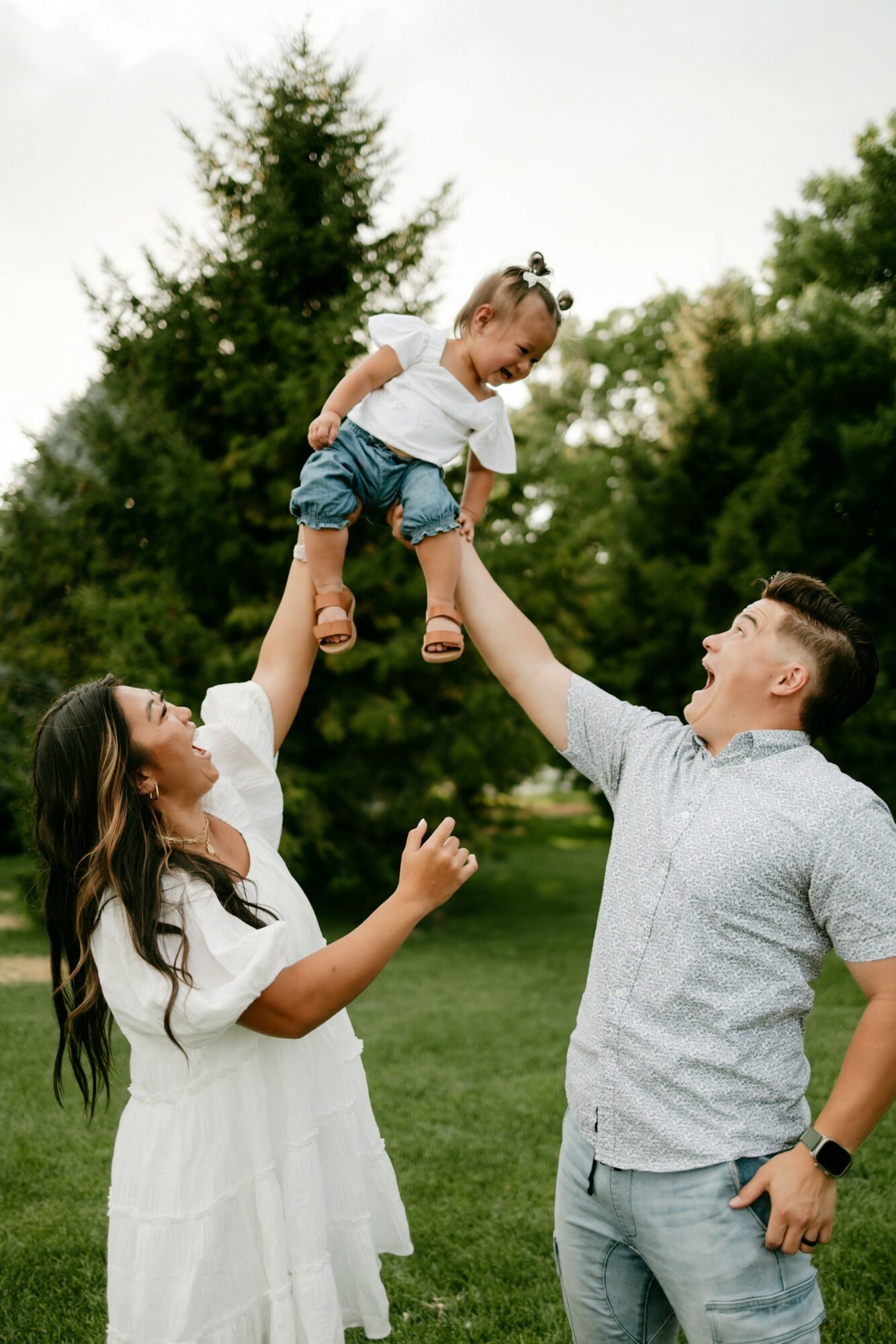 Family outdoors with a woman, man, and child, lifting the child in the air, trees in background, sunny day.