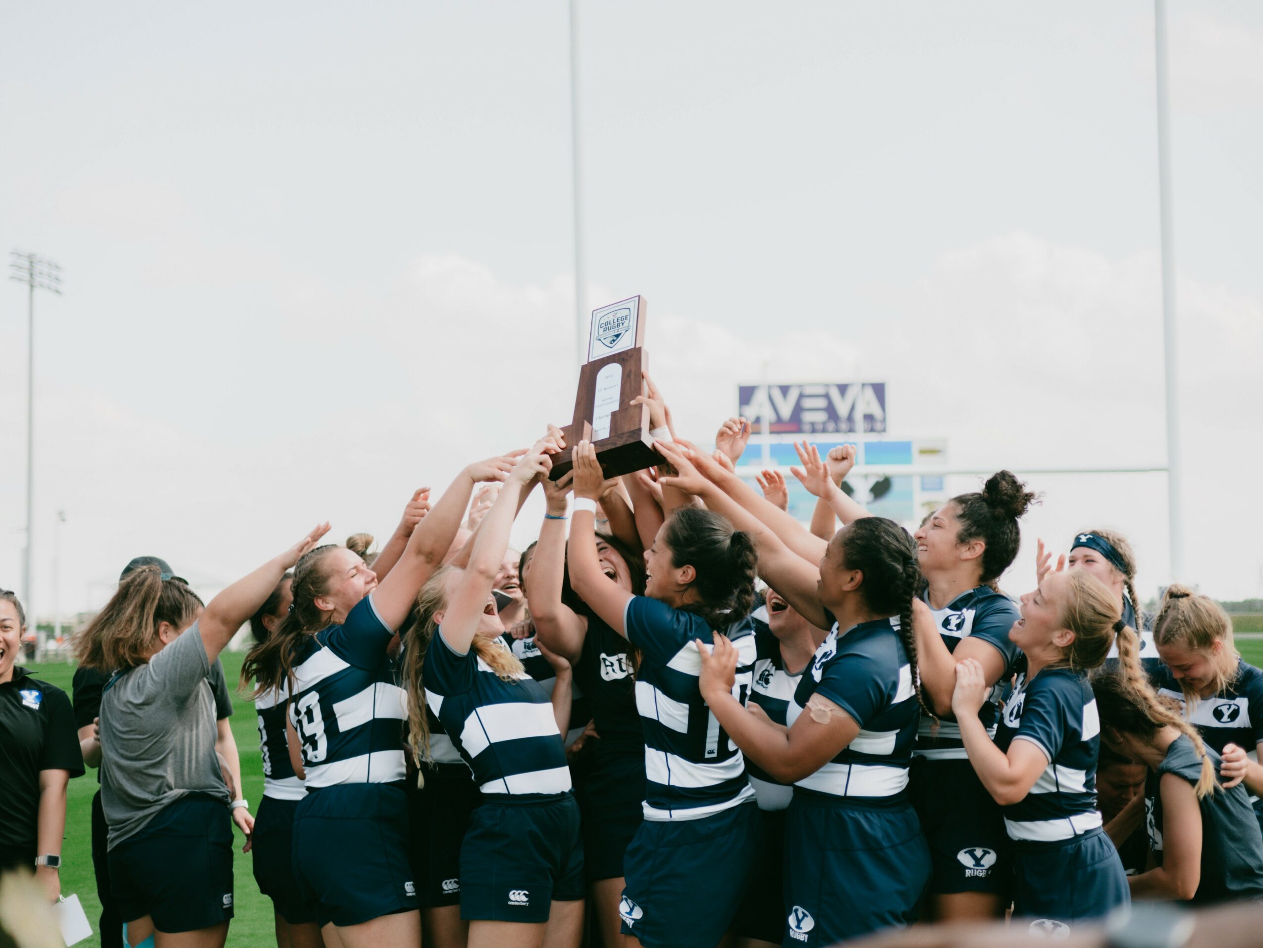 Group of female rugby players celebrating and holding a trophy on a field.