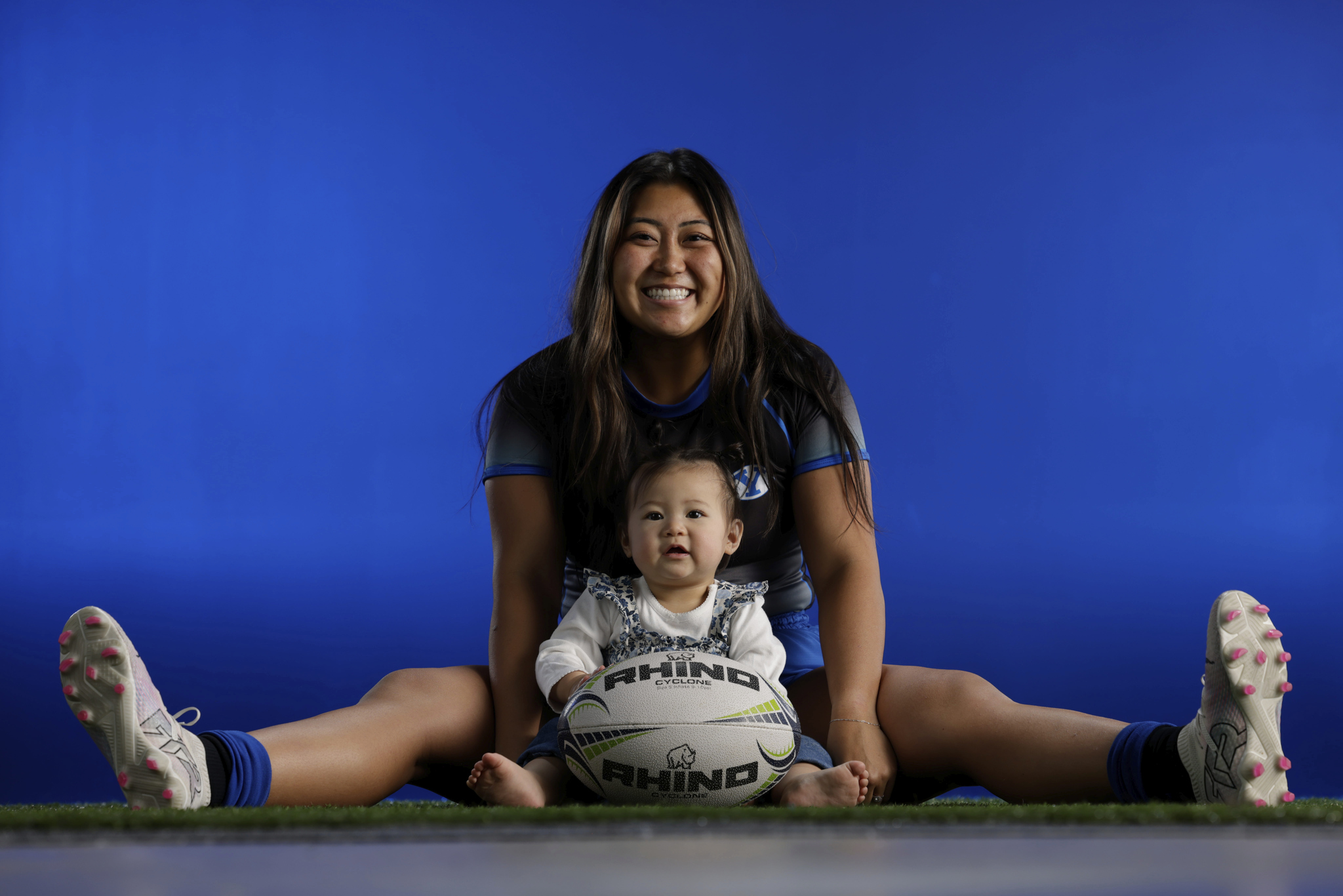 Woman and child sitting on the floor with legs spread, smiling, against a blue background, holding a soccer ball.