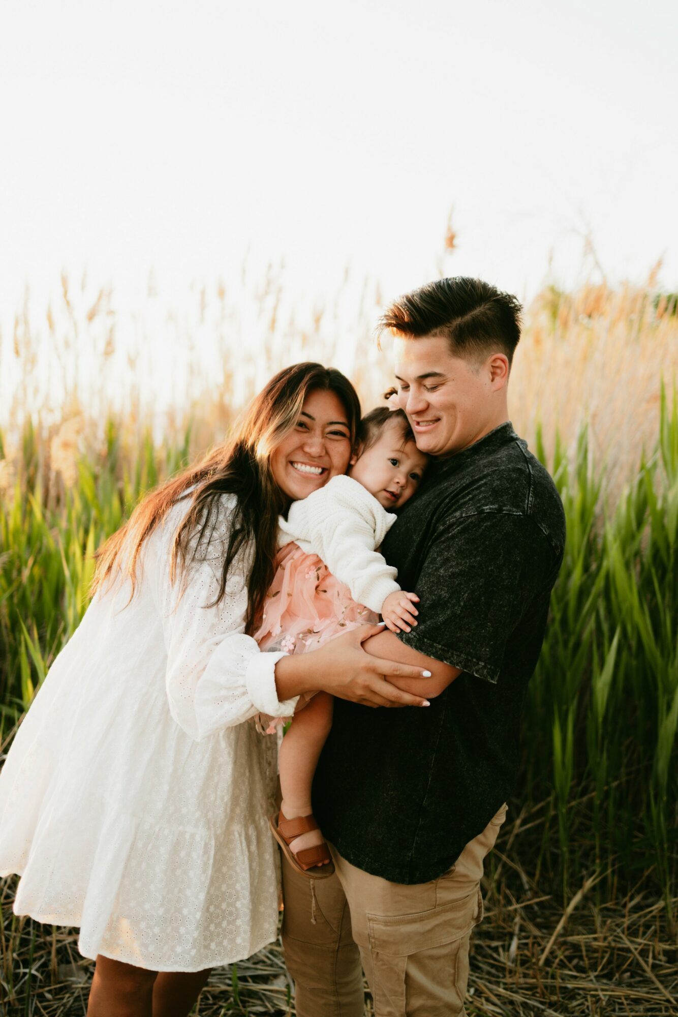 Family of four smiling outdoors in tall grass, embracing each other, sunny day, natural background.